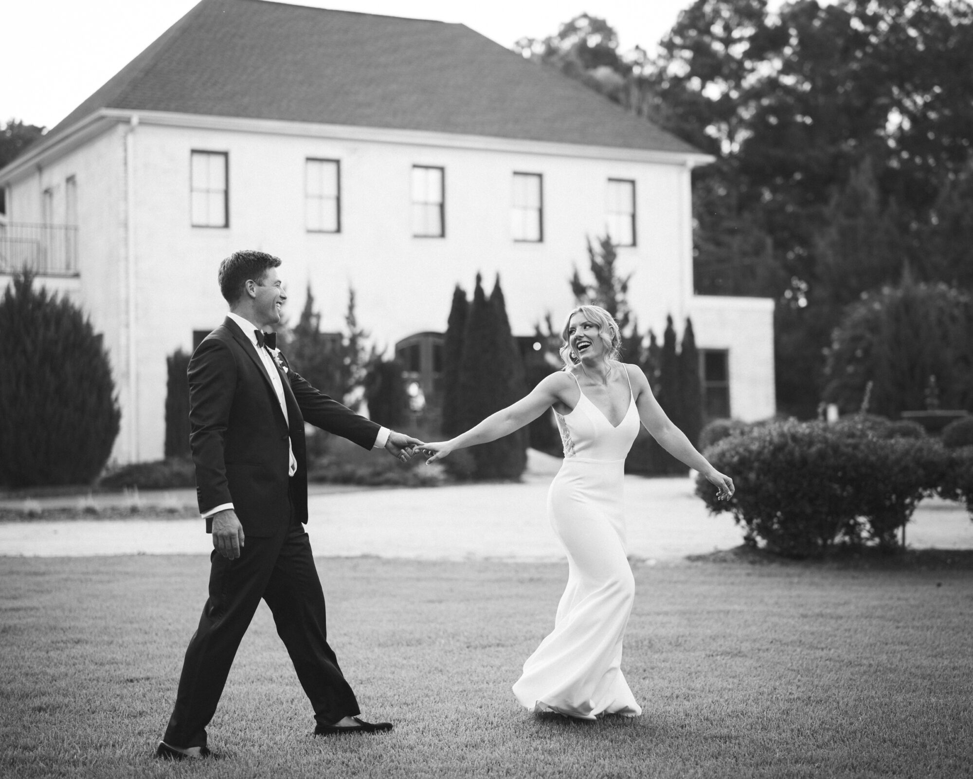 A couple in formal attire holds hands and smiles, capturing the joy of The Bradford Wedding, as they stroll on a grassy lawn before a large house with tall trees. The woman wears a white dress, and the man is in a dark suit. The image is captured beautifully in black and white.