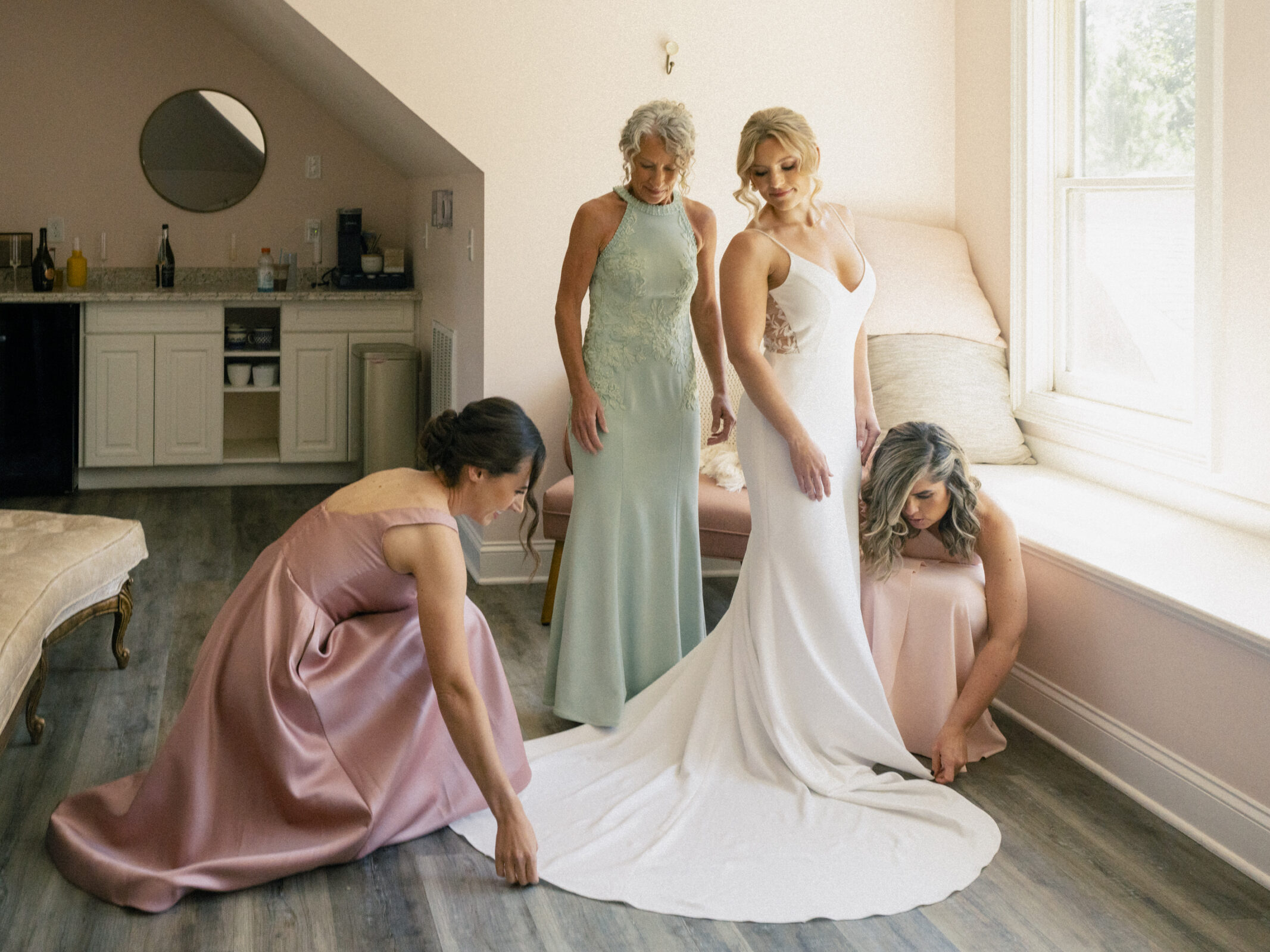 A bride in a white dress stands in a sunlit room at The Bradford Wedding venue as three women assist with her gown's train. One in pink and another in green adjust the dress, while the third in blue observes. They gather near a window that casts soft, natural light across the scene.