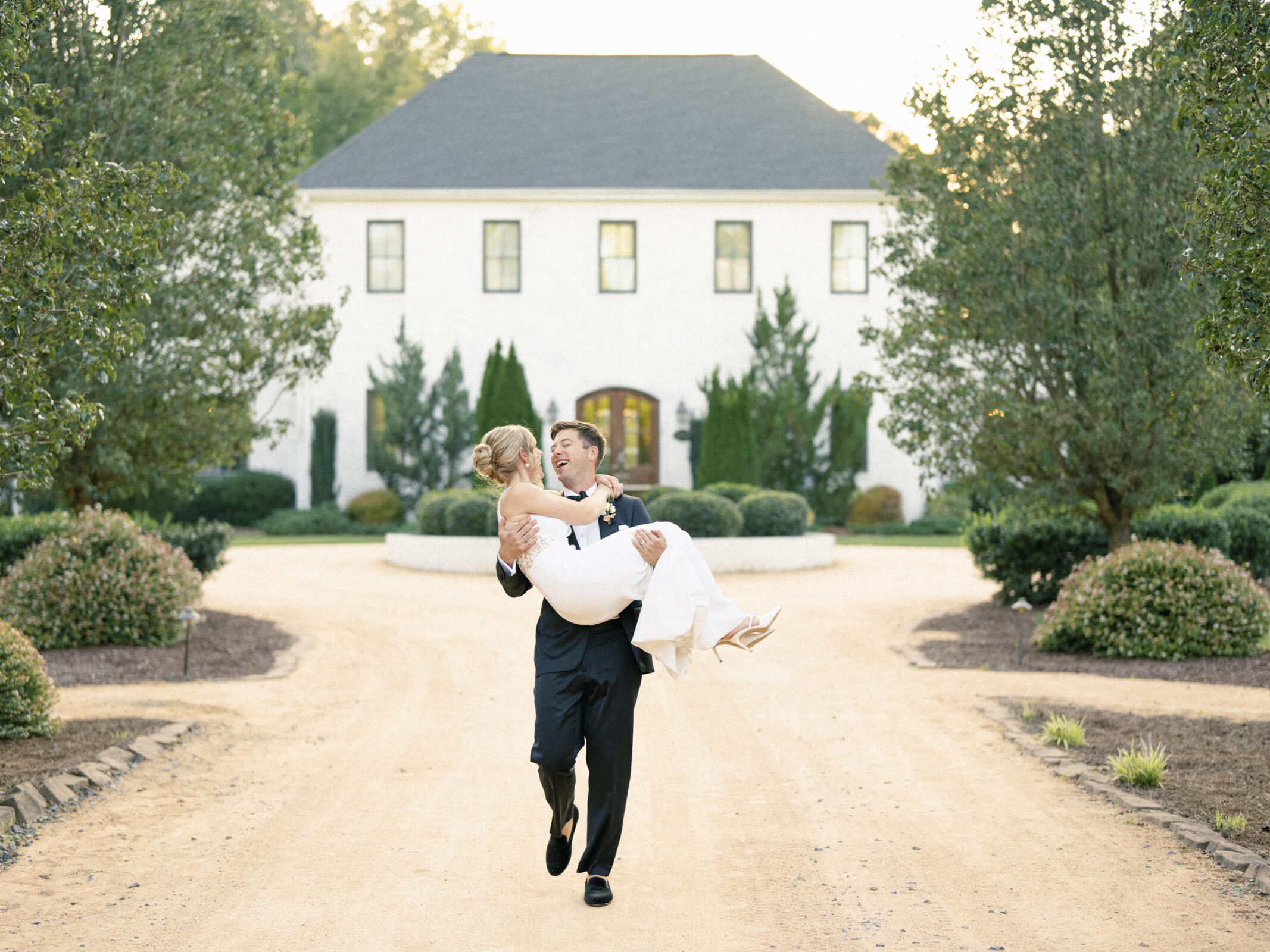 A groom joyfully carries the bride along a tree-lined path leading to The Bradford Wedding's grand white mansion. The bride wears a white dress, and both smile, surrounded by lush greenery under a clear sky, creating a romantic and elegant atmosphere.