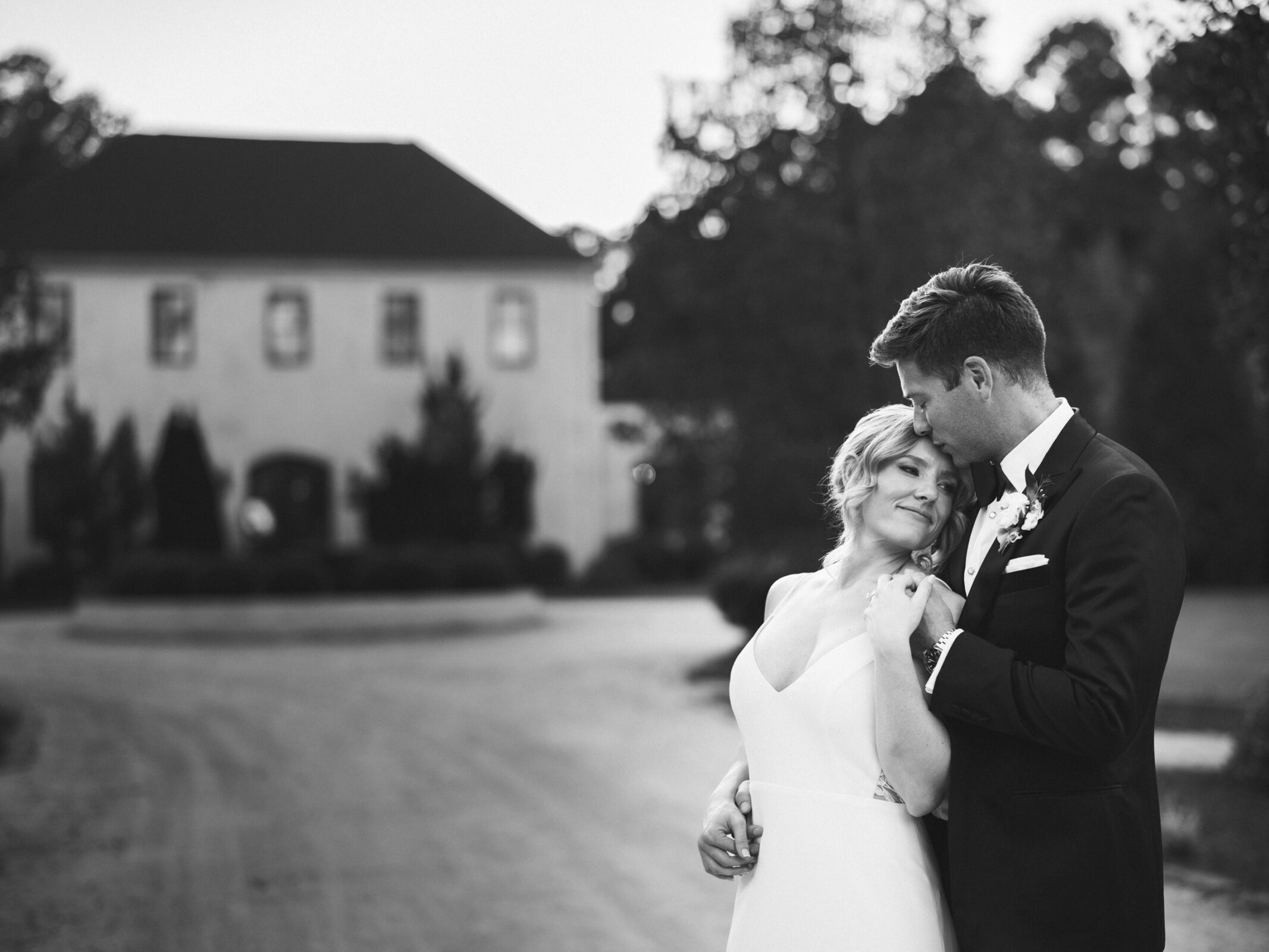 A black and white photo captures the essence of The Bradford Wedding. The couple embraces lovingly; the bride smiles while holding the groom's hand. They stand outside, framed by a large building and trees in the background, embodying timeless romance.