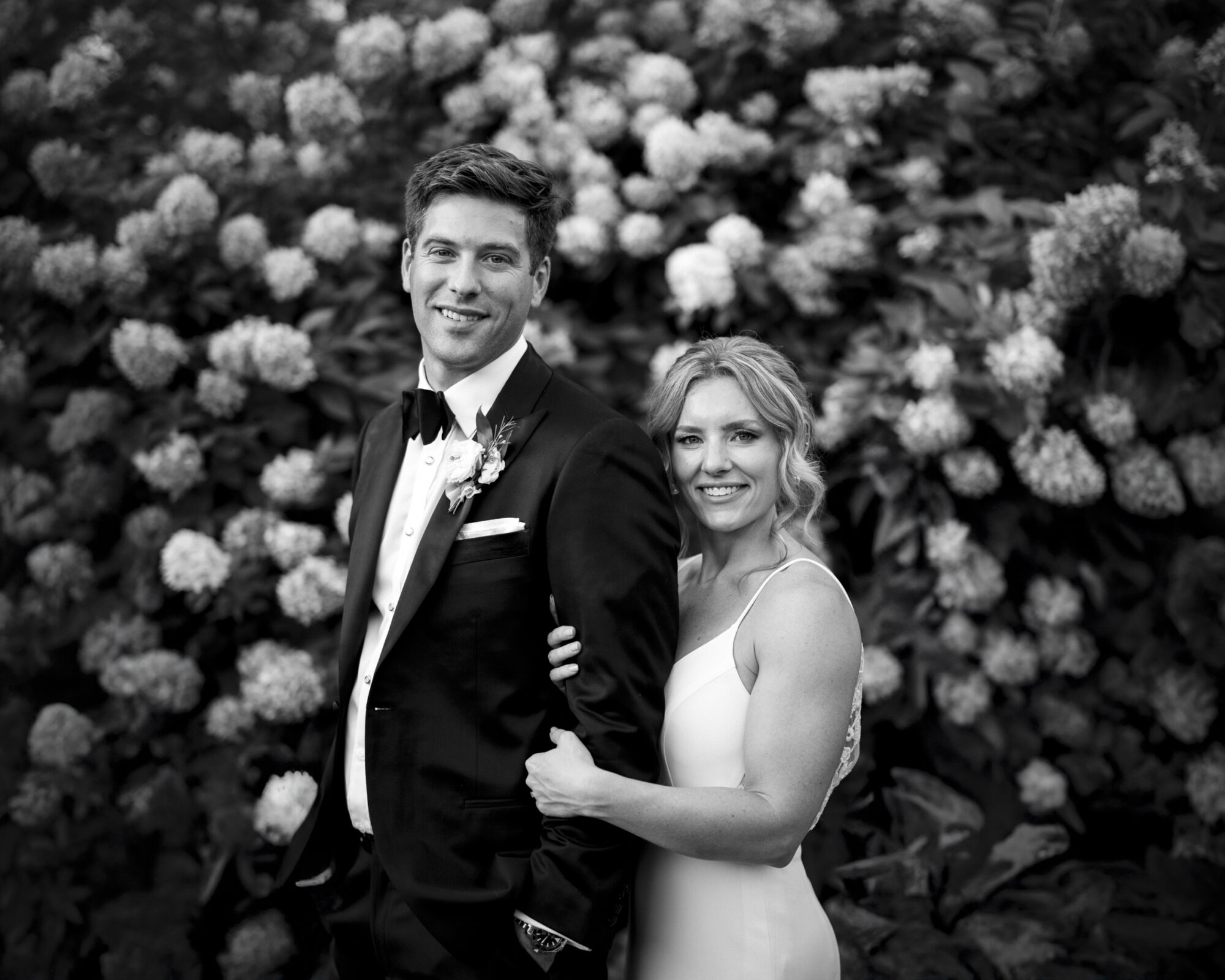 A black and white photo captures a couple at The Bradford Wedding, clad in formal attire. The man dons a suit and bow tie, as the woman stands behind him in a sleeveless dress, her hand resting on his shoulder. Both smile against the backdrop of blooming hydrangeas.