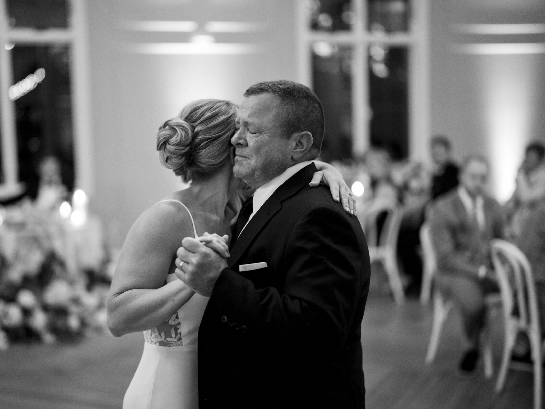 A black-and-white photo captures a touching moment at The Bradford Wedding, with a woman and an older man embracing while slow dancing in an elegant, softly lit venue. In the background, blurred guests sit at tables adorned with floral decorations.