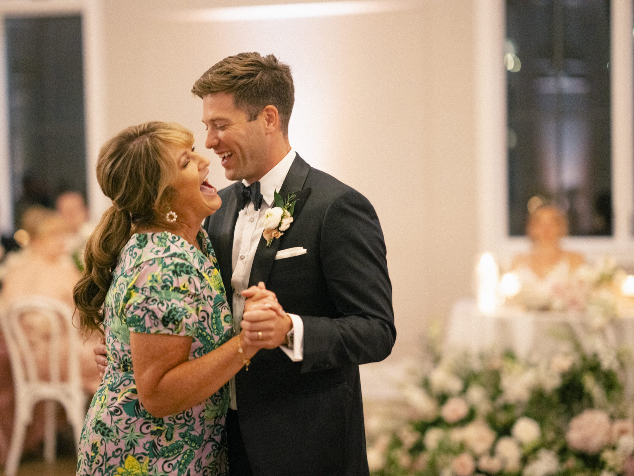 A joyful moment unfolds at The Bradford Wedding as a woman in a floral dress and a man in a tuxedo dance together. They smile at each other, surrounded by elegantly decorated tables and stunning floral arrangements, perfectly capturing the beauty of the celebration.