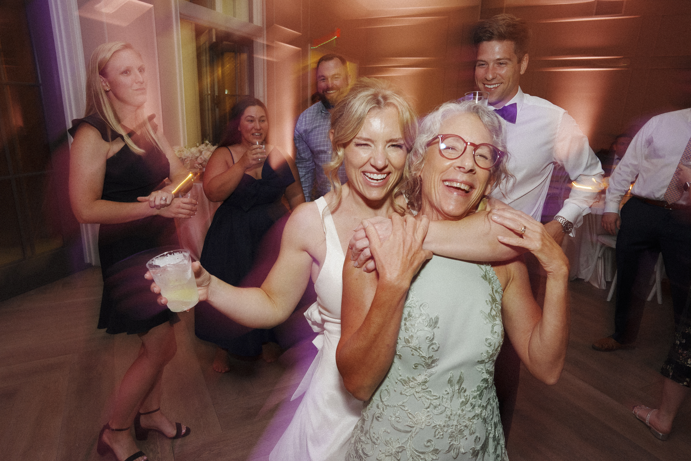 At The Bradford Wedding, a joyful bride in a white dress embraces an older woman in a light green dress on the dance floor. Surrounded by smiling guests, including a woman holding a drink, the setting is lively and festive.