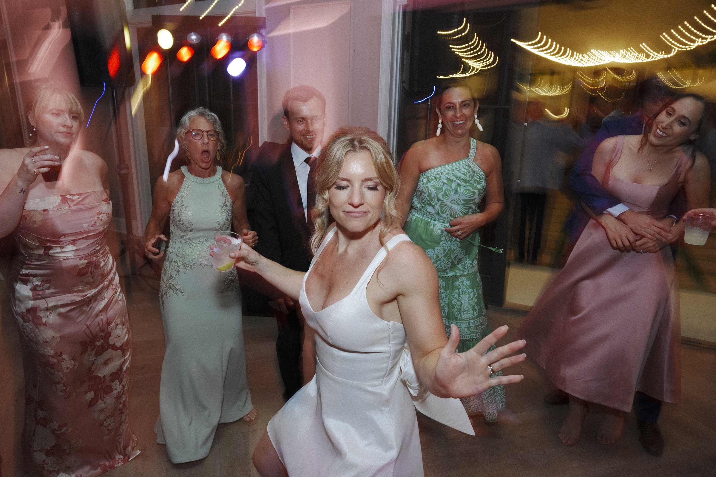 At The Bradford Wedding, a group of people dances enthusiastically at the party. In the foreground, a woman in a white dress strikes a playful pose while others around her smile and watch. Colorful lights in the background create a festive atmosphere.