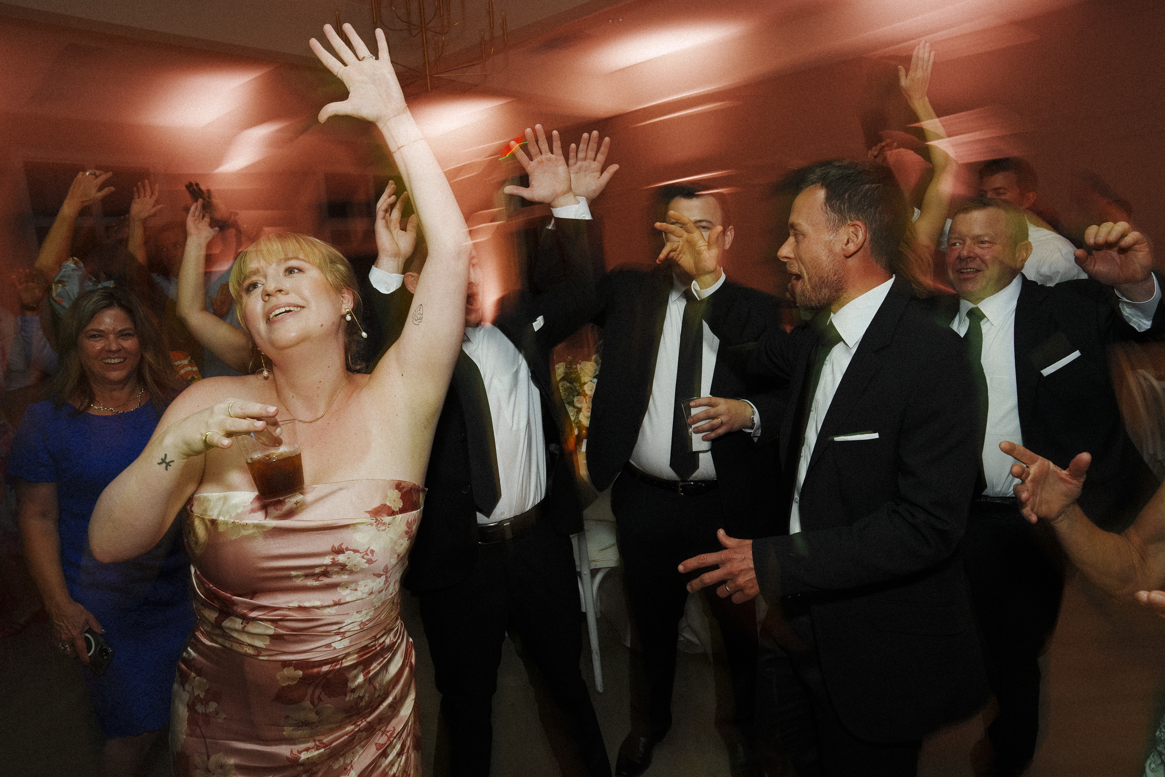 At The Bradford Wedding, a joyful group of people dances at the celebration. In the foreground, a woman in a floral dress holds a drink and raises her arm. Men in suits around her join in enthusiastically. The atmosphere is lively and festive.