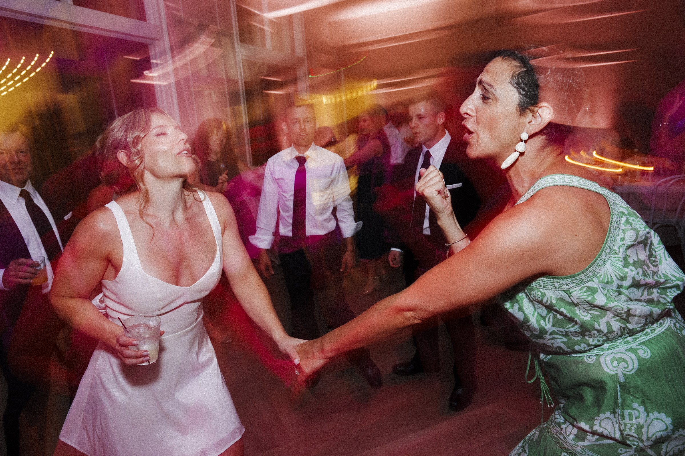 At The Bradford Wedding, two women dance energetically in a lively room with colorful lights. One wears a white dress and holds a drink, while the other dazzles in green. Several guests mingle in the background, adding to the festive atmosphere.