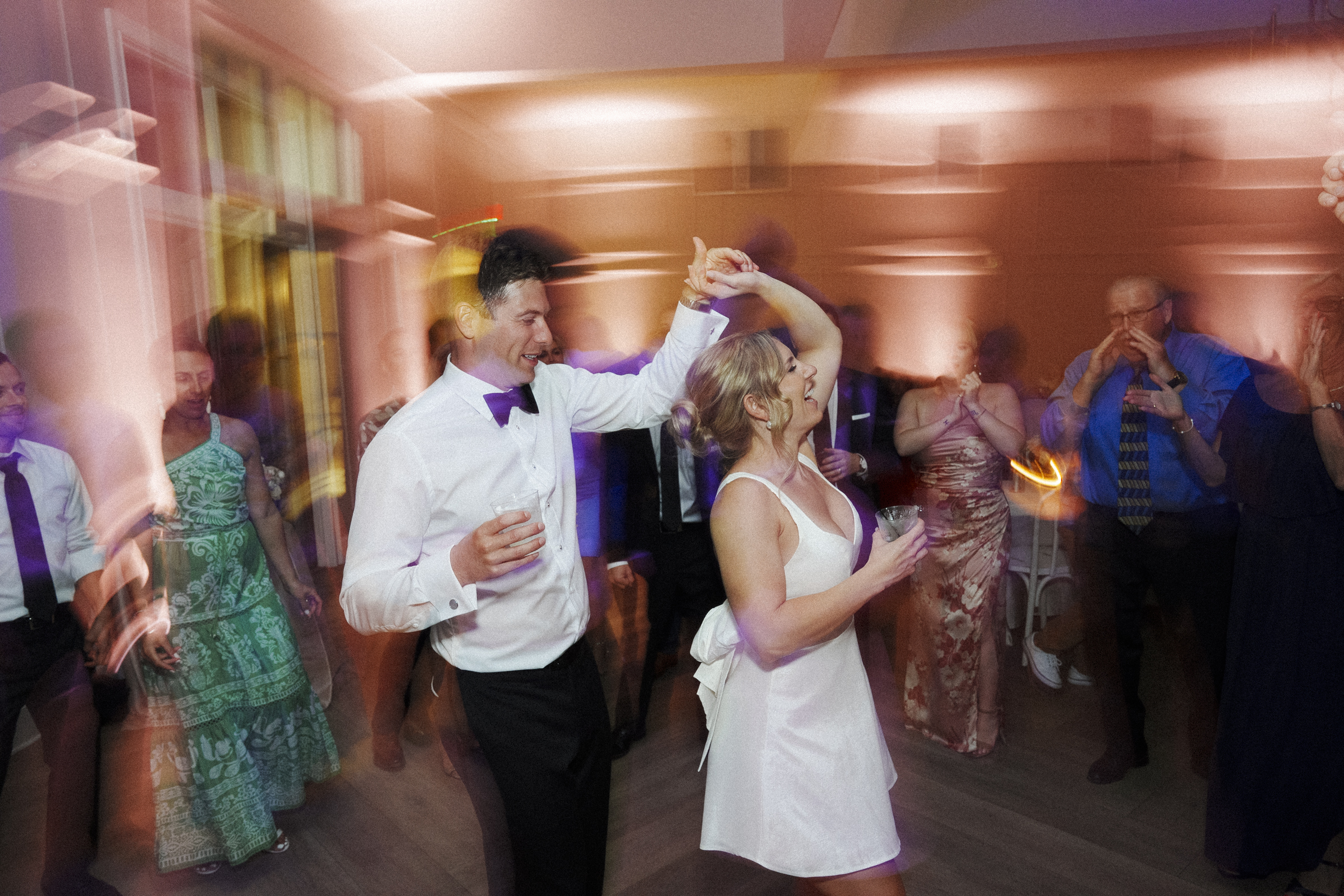 At The Bradford Wedding, a couple dances joyfully at the lively party. The man wears a white shirt and bow tie while the woman is in a dazzling white dress. Blurred lights and other guests dancing in the background create a vibrant, festive atmosphere.