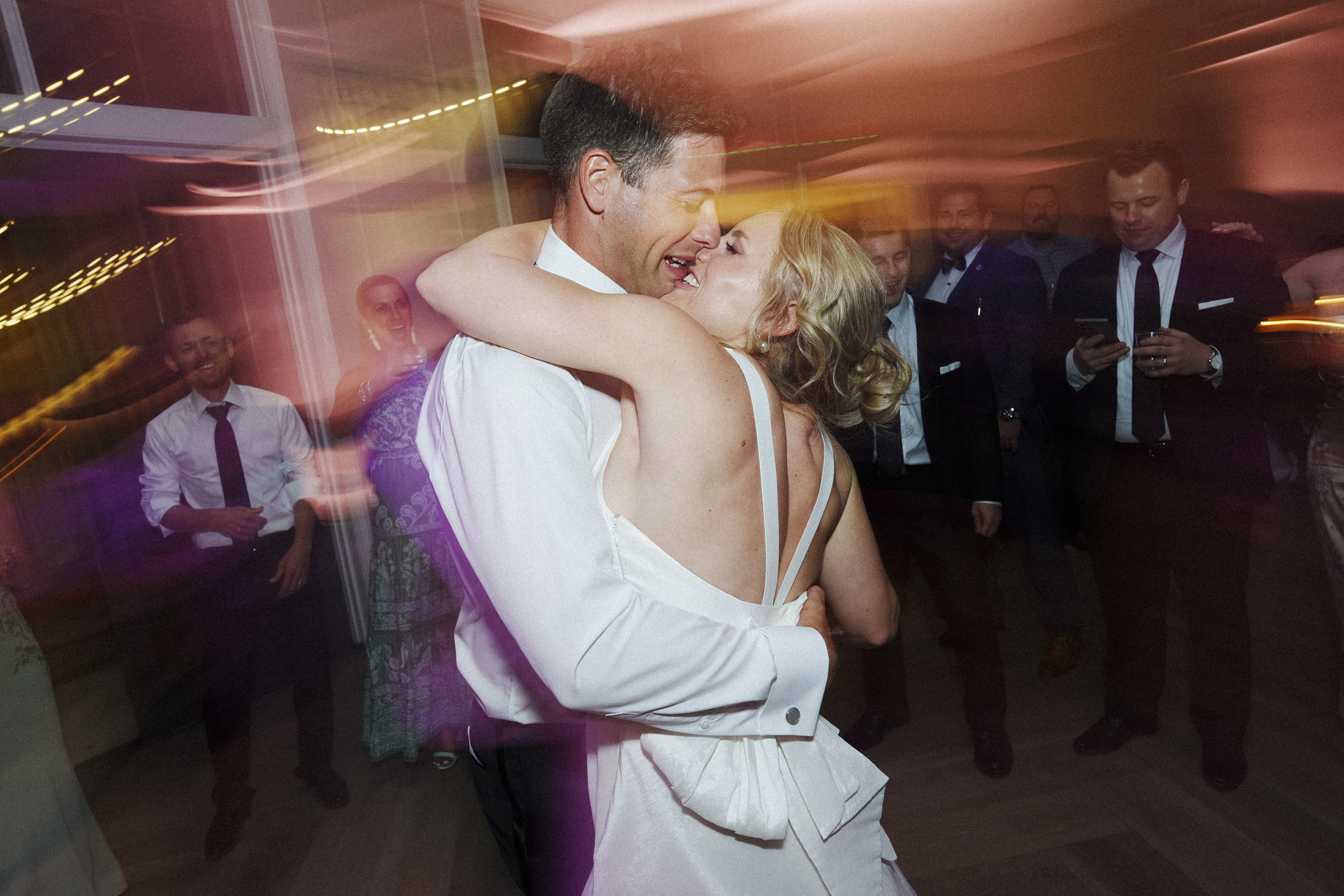 At The Bradford Wedding, a couple shares a joyful dance at the reception. The bride in a white dress embraces the groom as guests, dressed formally, watch and smile in the dimly lit room with warm lighting. Movement blur adds a dynamic feel to this enchanting scene.