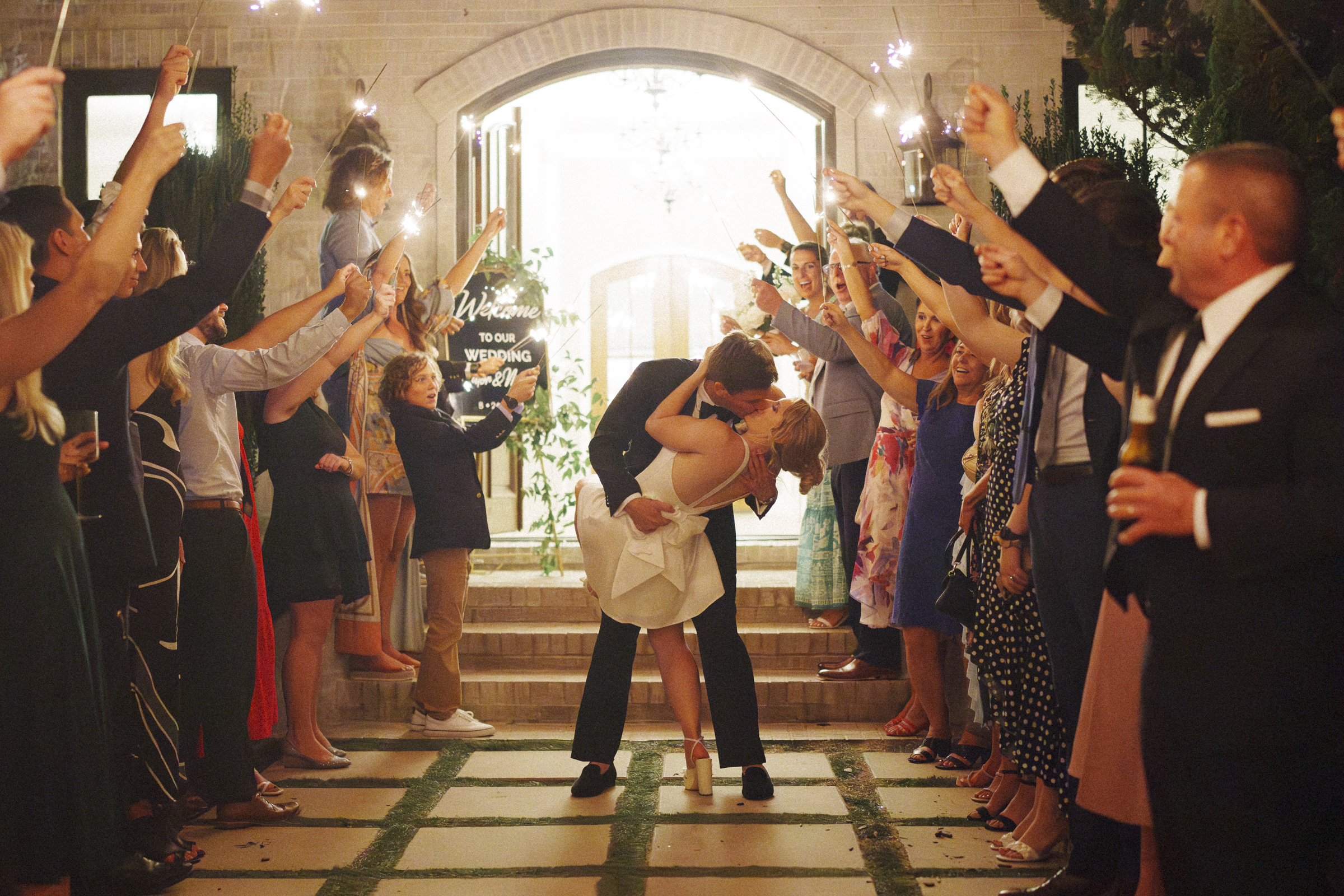 A couple kisses passionately under sparklers during their Bradford Wedding exit. Guests stand on either side, celebrating and cheering them on. The bride's white dress and the groom's suit contrast against the warmly lit scene.