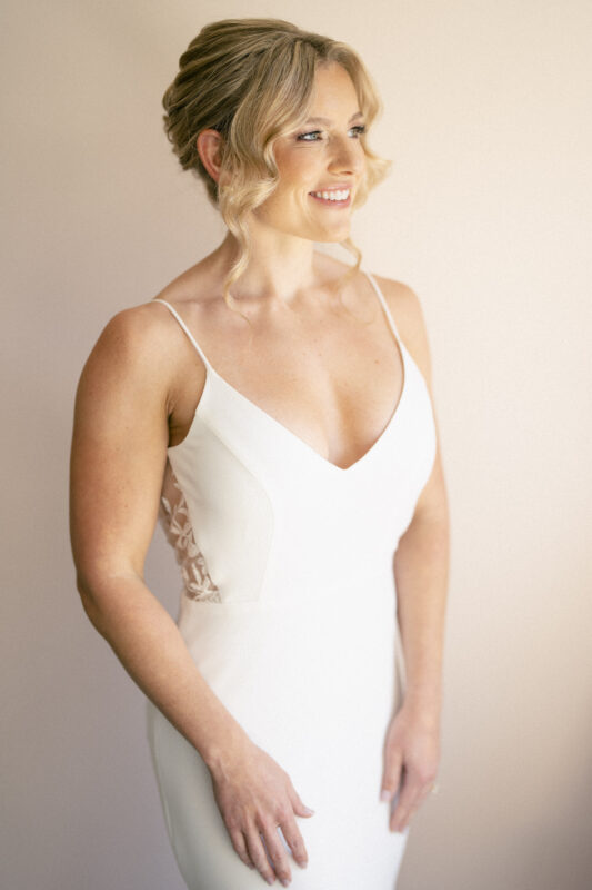 A woman in a white wedding dress from The Bradford Wedding collection stands smiling. Her hair, styled in an updo with loose curls, frames her face as she gazes slightly to the side against a plain, soft backdrop.