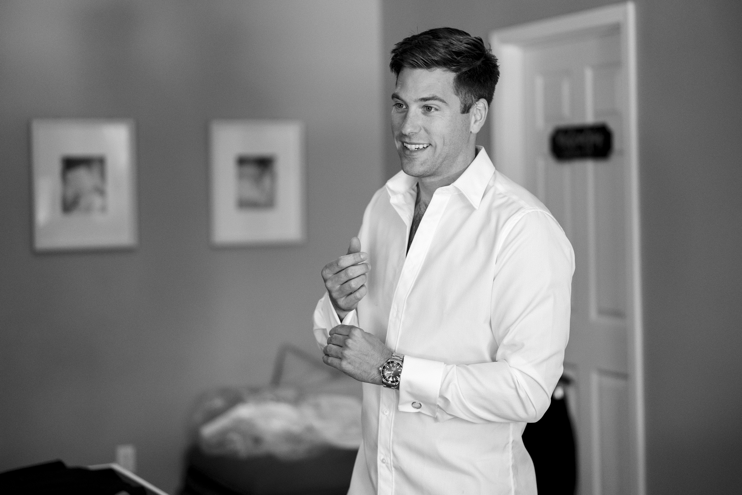 A man in a white dress shirt stands in a softly focused room, adjusting his sleeve and smiling. Framed pictures adorn the walls, hinting at cherished memories from The Bradford Wedding, while a door lingers quietly in the background. The timeless image captures elegance in black and white.