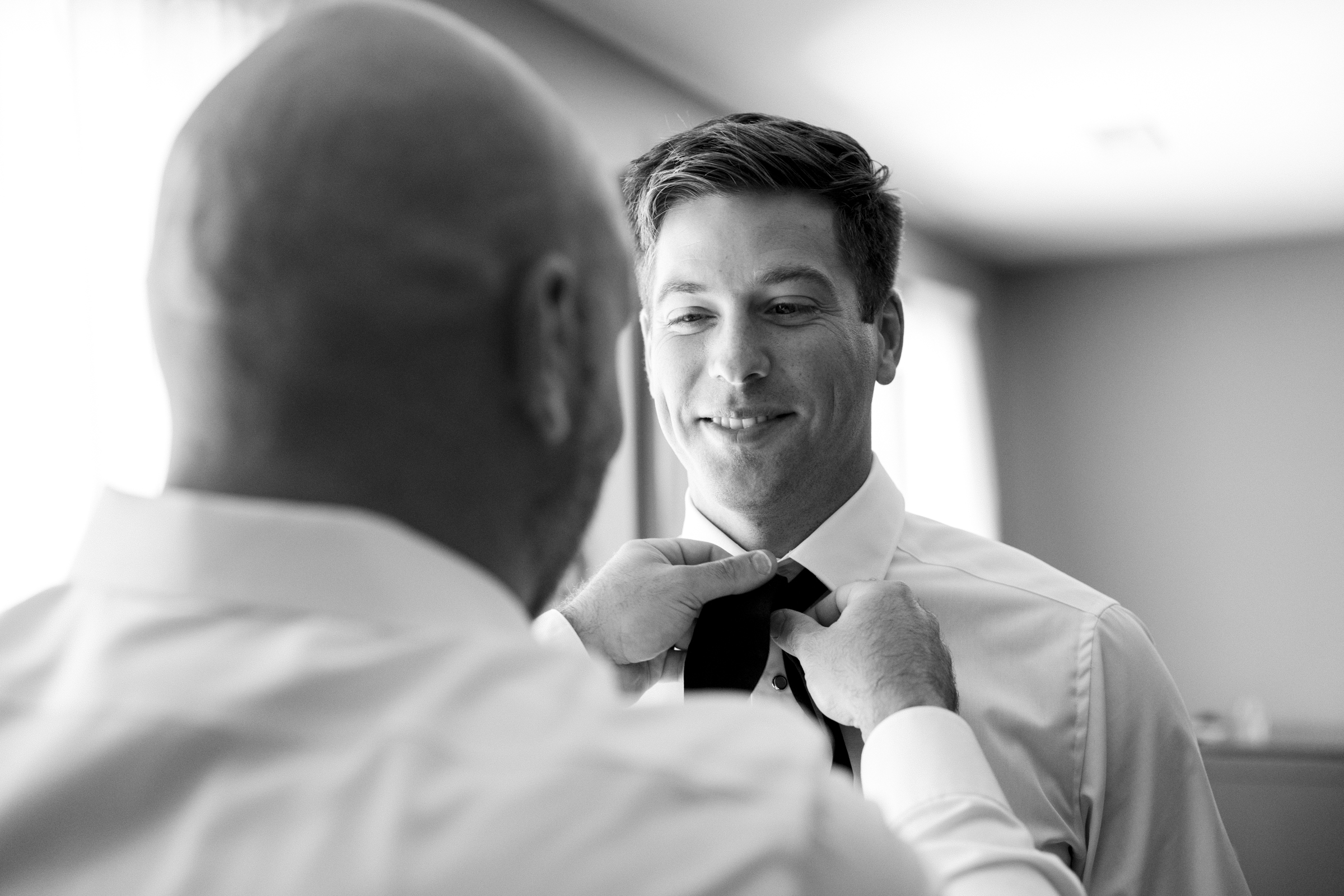 A man smiling as another adjusts his tie, both in dress shirts, illuminated by natural light streaming through a window—a charming moment captured at The Bradford Wedding.