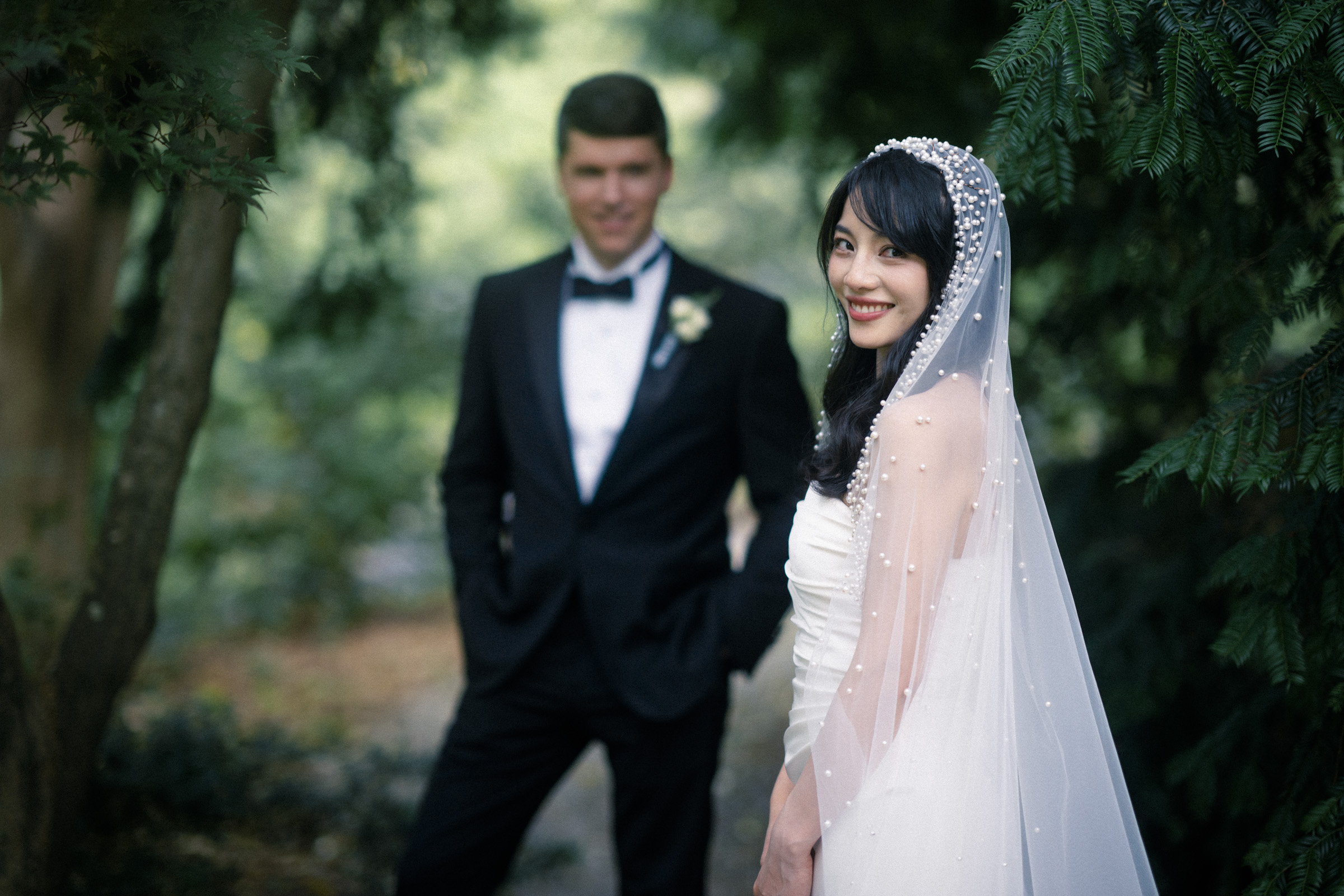 A bride in a white gown and veil smiles at the camera, standing in a lush garden. In the background, a groom in a black tuxedo stands, slightly out of focus. Trees and greenery surround the couple, creating a serene atmosphere.