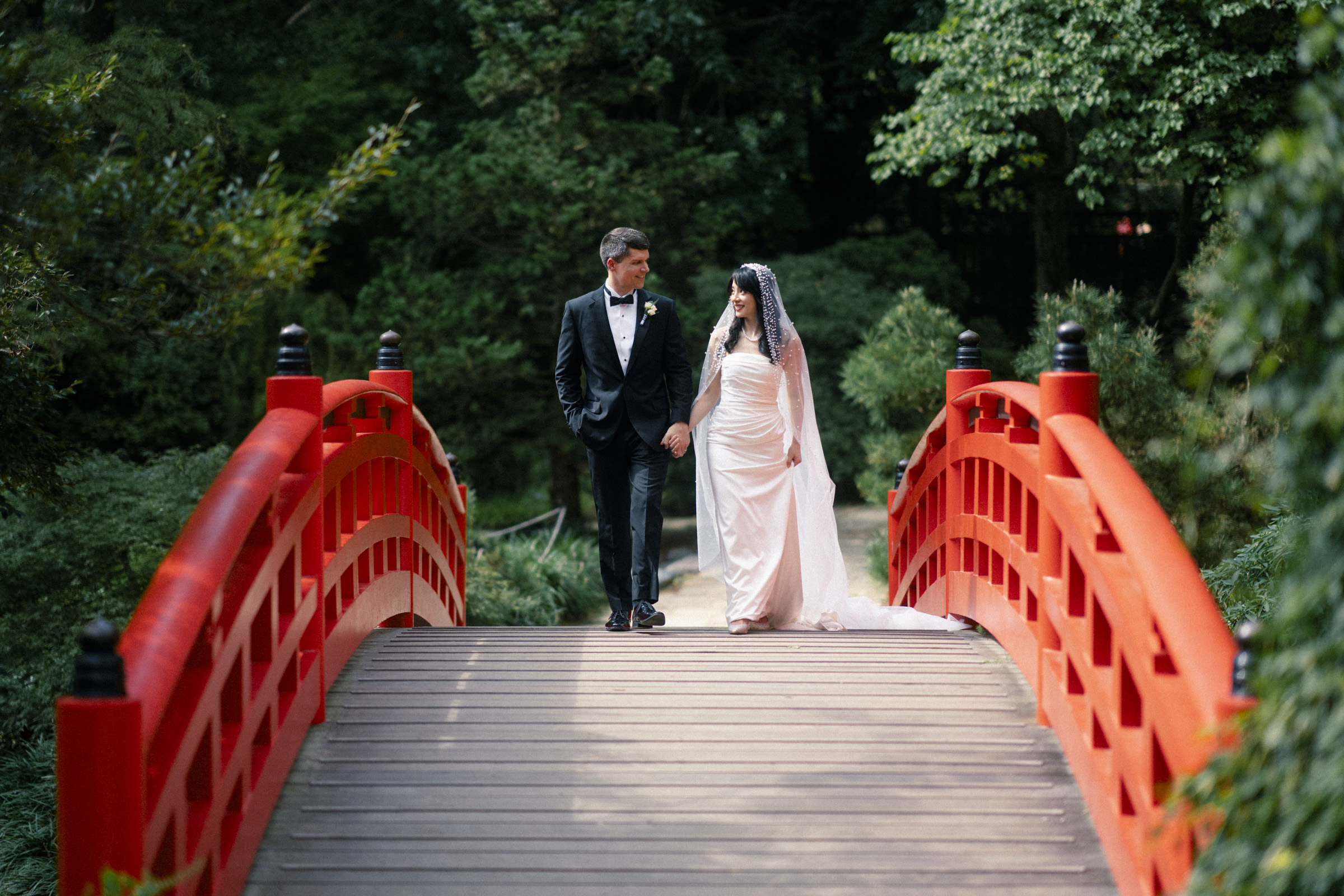 A bride and groom walk hand in hand across a red arched bridge in a lush garden, surrounded by green trees. The bride wears a white gown and veil, while the groom is in a black suit. The scene is serene and romantic.