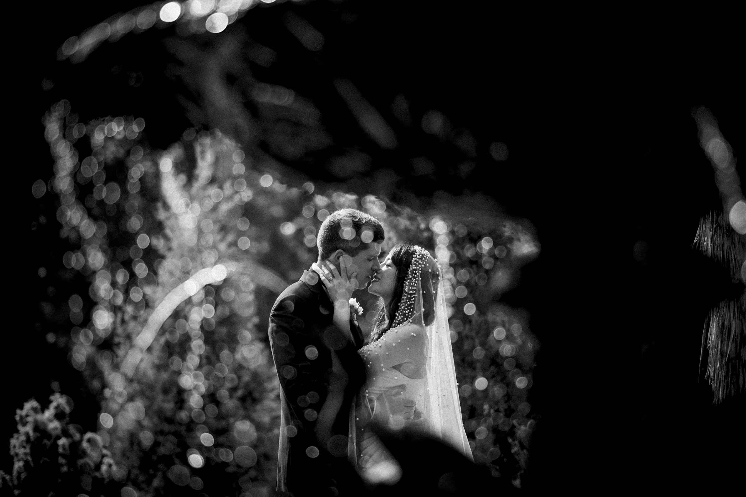 A black-and-white photograph captures a bride and groom embracing under dappled sunlight at the Duke Chapel wedding. The blurred background highlights their tender moment as the bride's veil gently dances, and their expressions reveal a profound emotional connection.