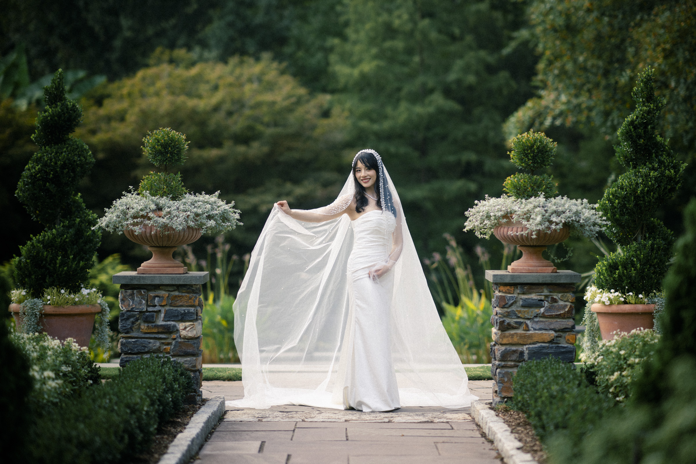 A bride in a white wedding dress and veil stands outdoors in a lush green garden. She is smiling and holding the sides of her veil, surrounded by neatly trimmed hedges and large potted plants on stone pedestals.