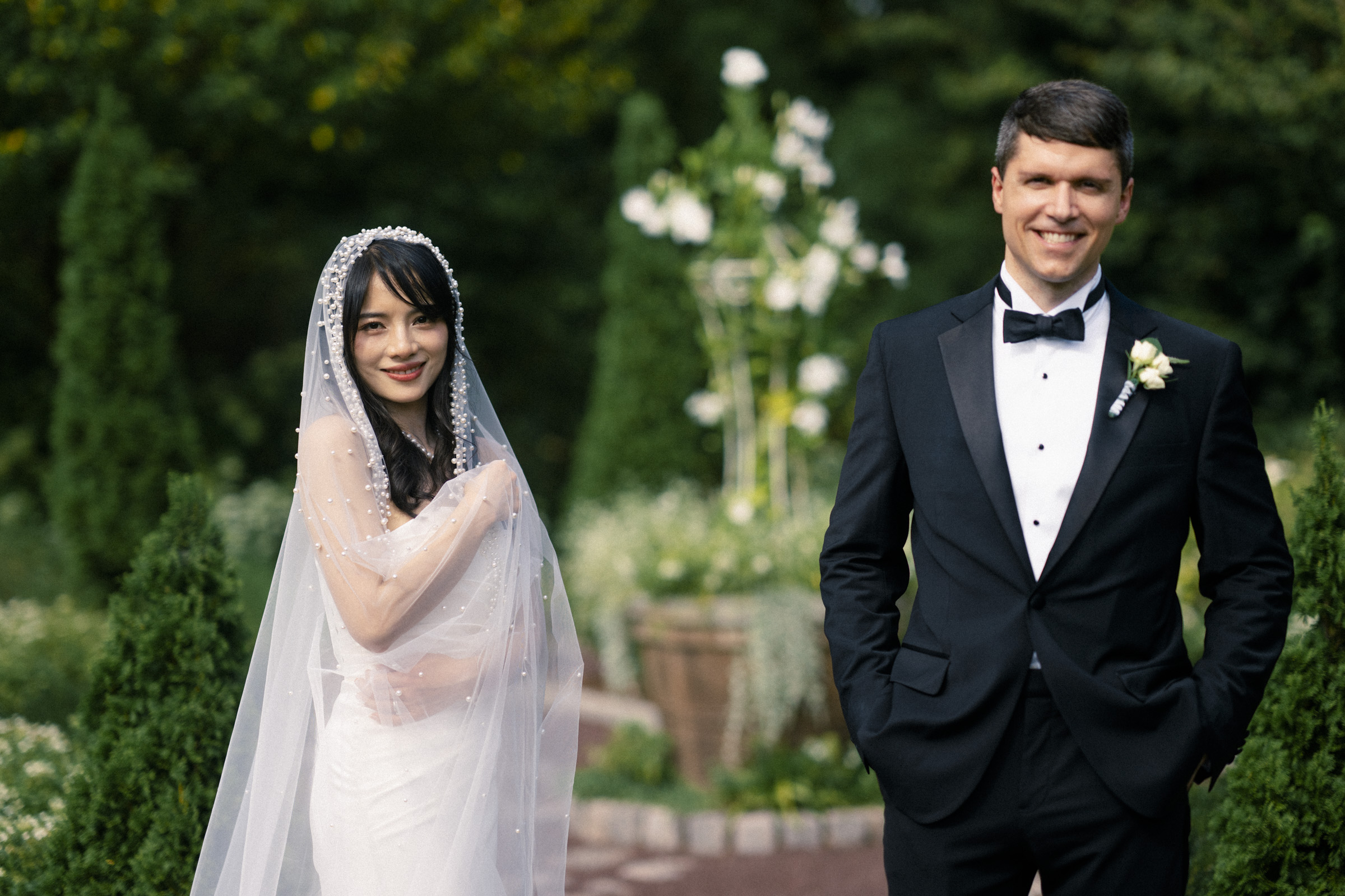 A bride in a white gown with a beaded veil stands beside a groom in a black tuxedo, both smiling. They are outdoors in a garden setting with lush greenery and white flowers in the background.