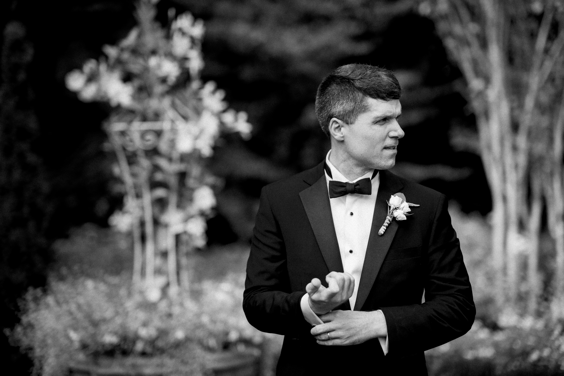 A man in a tuxedo stands in a garden setting with blurred floral decorations in the background. He is adjusting his cufflink and looking to the side. The image is in black and white.