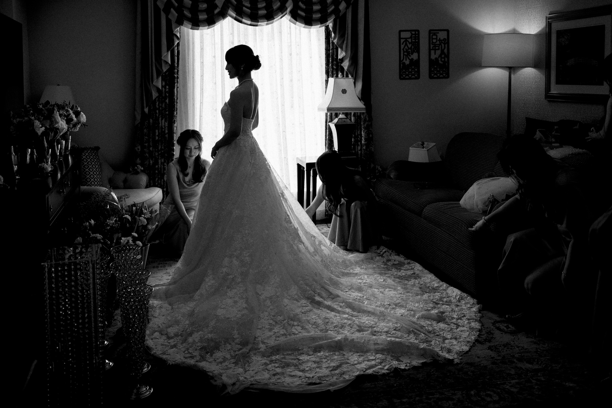 A black and white photo of a bride in a long, lacy wedding gown standing in a dimly lit room. Two bridesmaids adjust the dress near her feet. The room has elegant furnishings and a window with sheer curtains in the background.