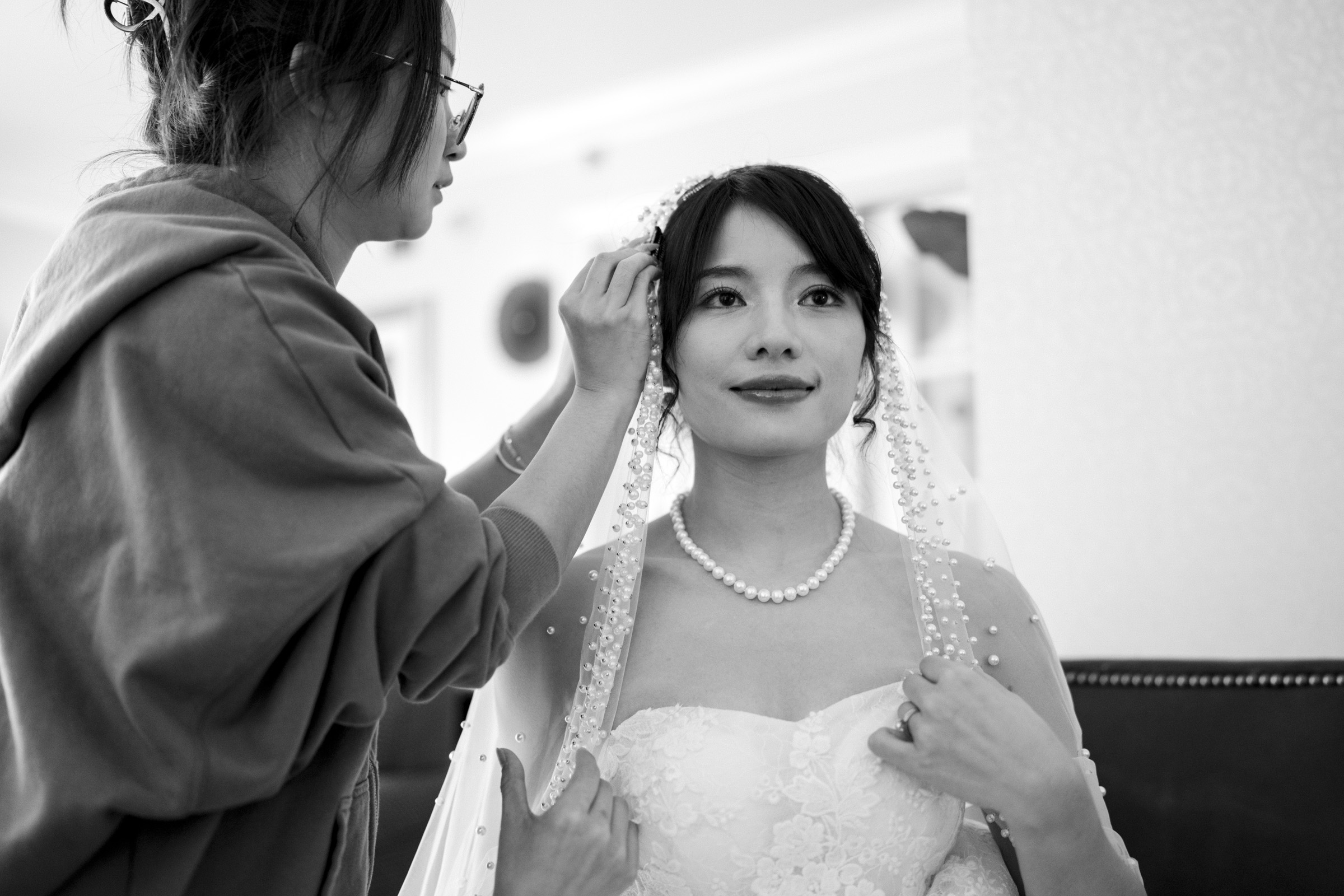 A bride wearing a strapless lace wedding dress and pearl necklace is getting her hair adjusted by a stylist. The stylist is focused, adjusting the brides veil. The image is in black and white, capturing an intimate bridal preparation moment.