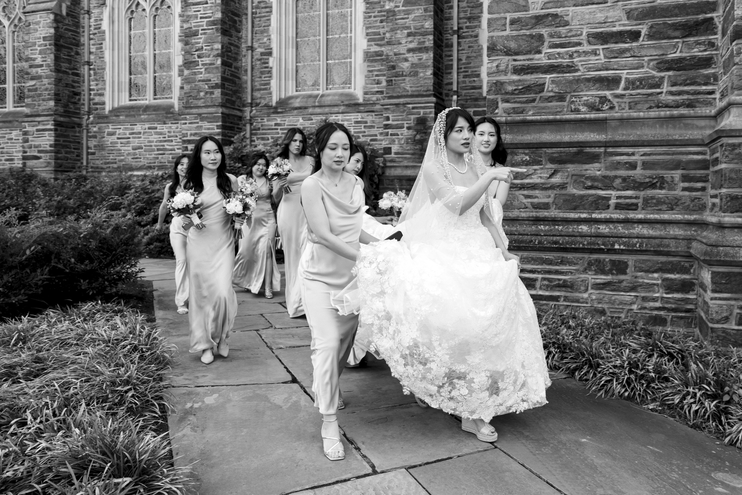 A black and white image of a bride in a lace gown walking with bridesmaids in dresses outside a stone church. The women hold bouquets and assist with the brides dress as they move along a paved path.