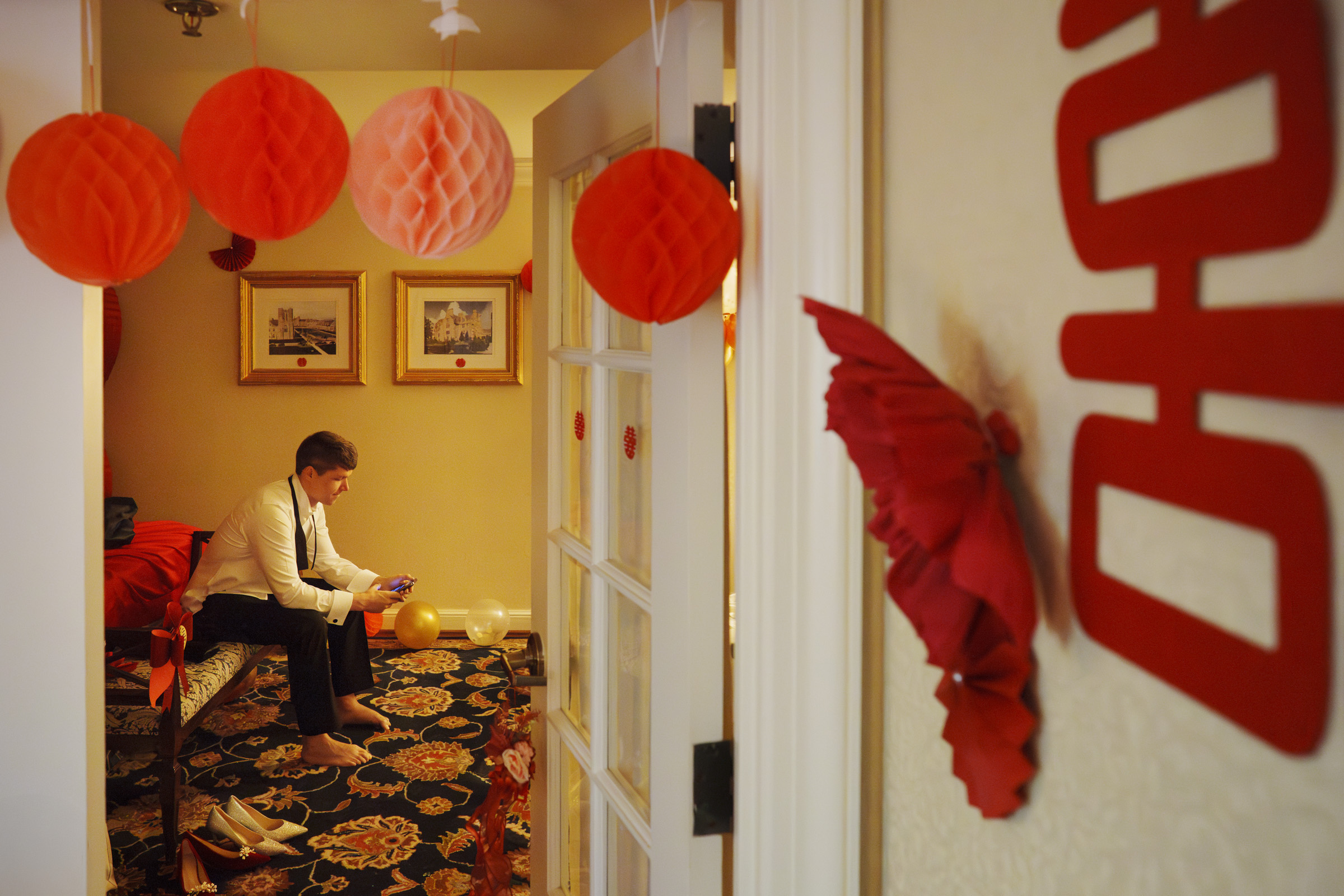 A person in formal attire sits on a bed in a room decorated with red and orange hanging paper lanterns and wall decorations. The room has framed artwork and patterned carpet. They are using a smartphone.