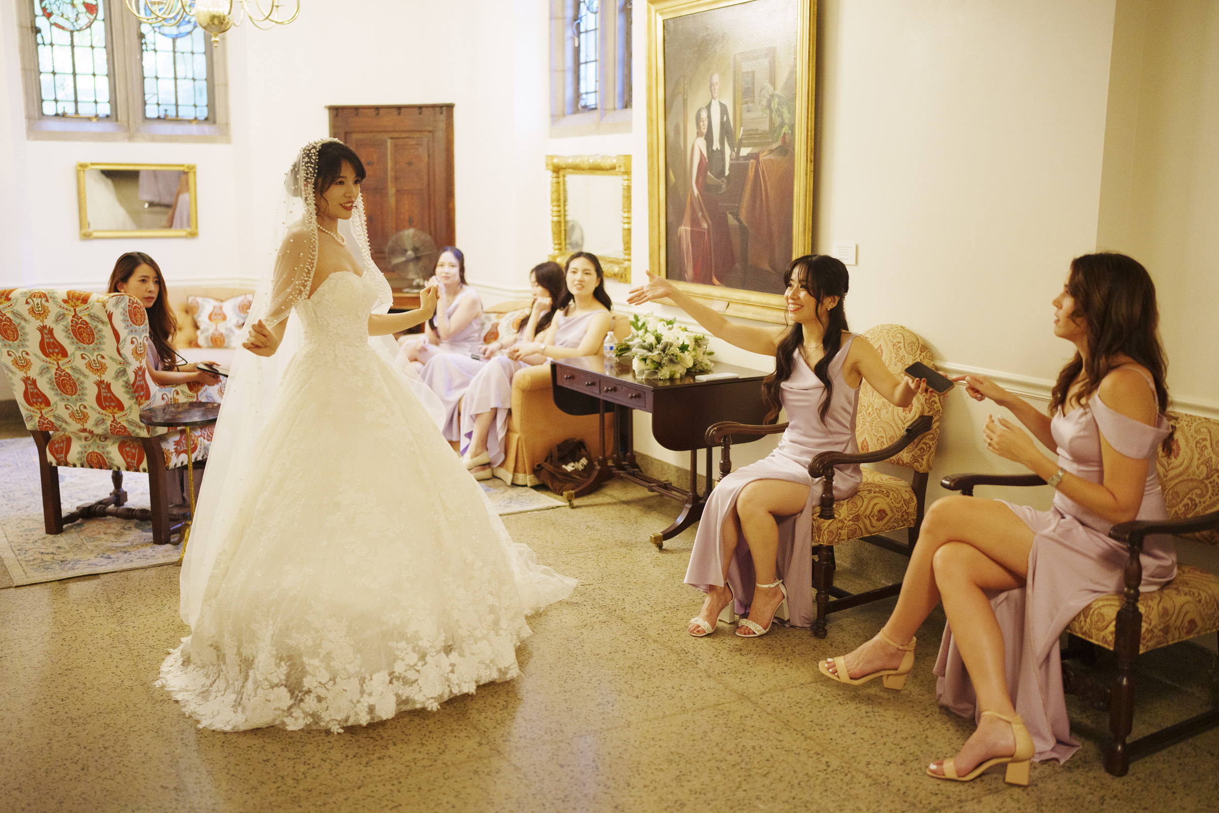 A bride in a white gown stands joyously, surrounded by bridesmaids in lavender dresses sitting on sofas. The room is elegantly decorated, featuring a chandelier, large windows, and a framed painting on the wall. Everyone appears happy and excited.