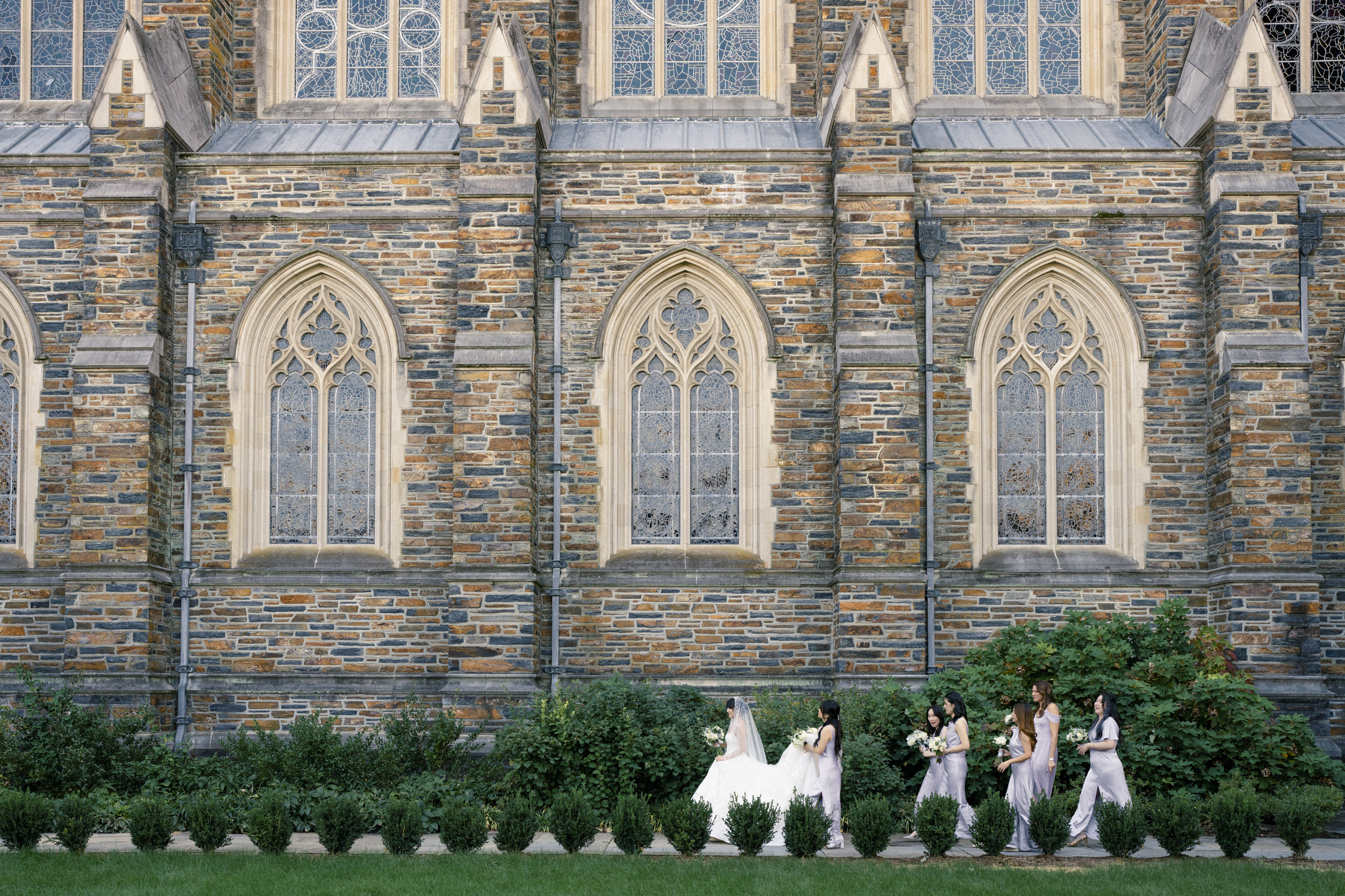 A bride in a white gown walks along the side of a large stone church with arched windows, accompanied by bridesmaids in taupe dresses carrying flowers. The scene is framed by lush green bushes and grass.