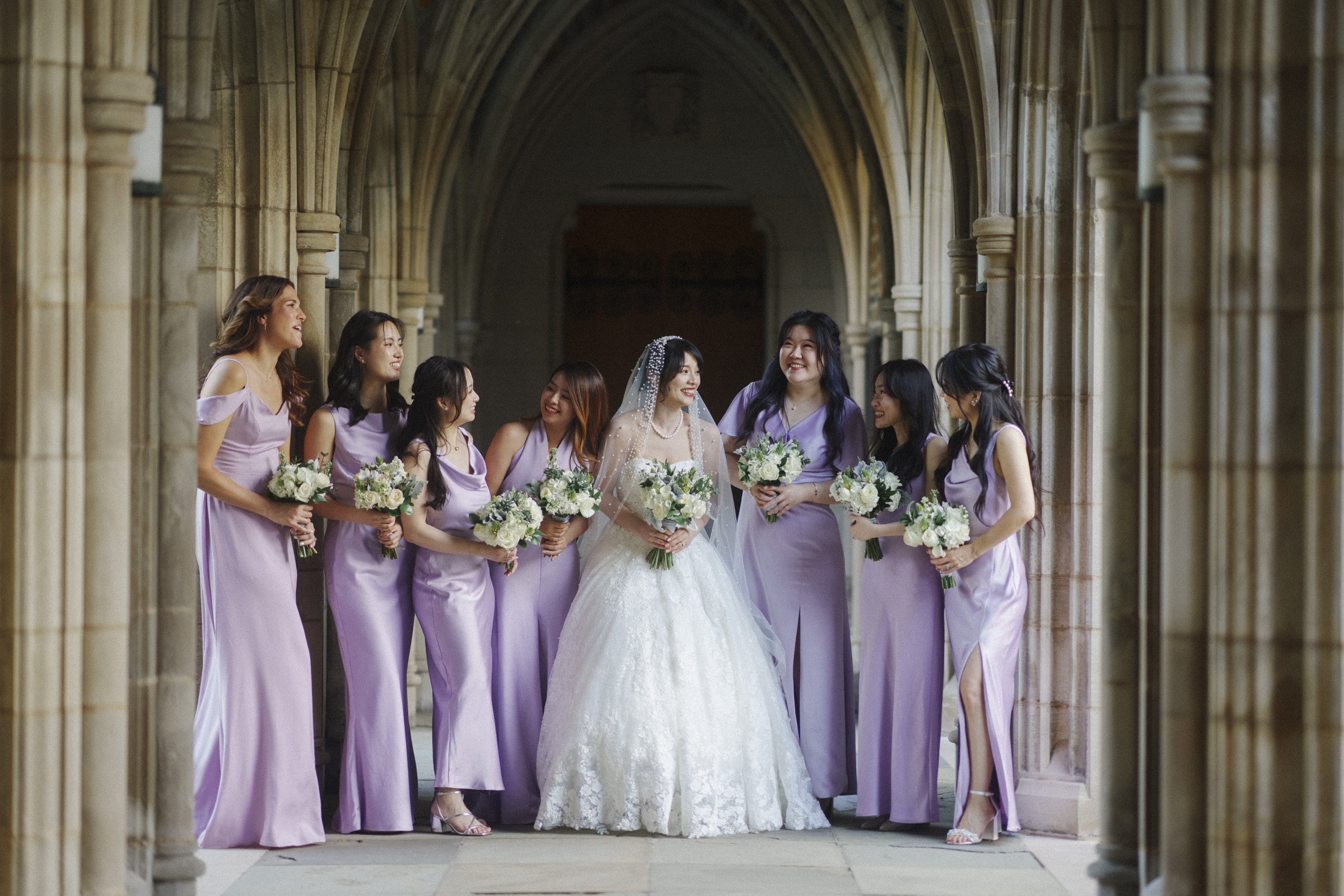 A bride in a white gown stands in an ornate archway with six bridesmaids in lavender dresses. They are all holding bouquets, smiling, and looking at each other, creating a joyful scene.