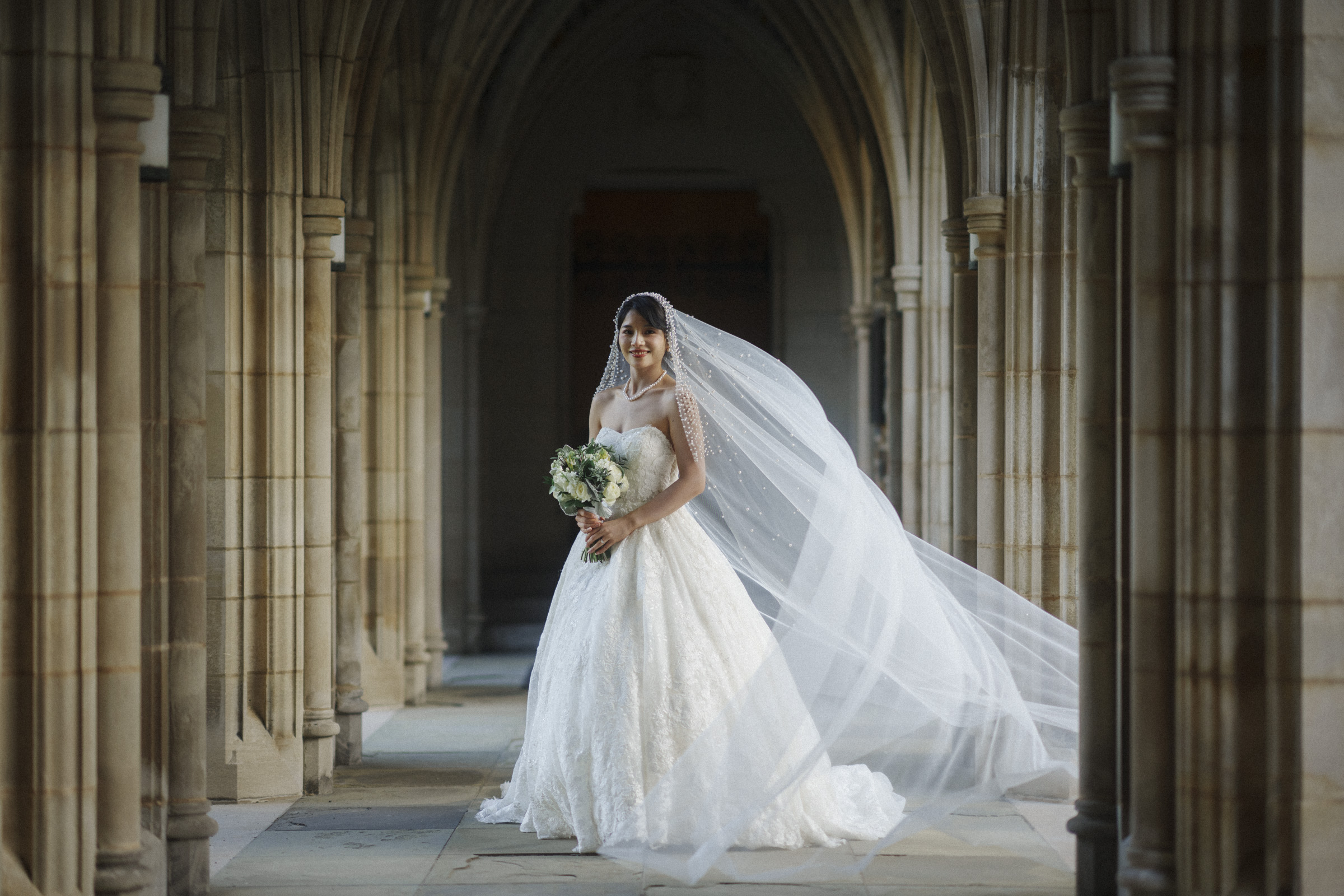 A bride stands in a stone archway, wearing an elegant white wedding gown with a long veil flowing behind her. She holds a bouquet of flowers and smiles softly, with natural light illuminating the scene.