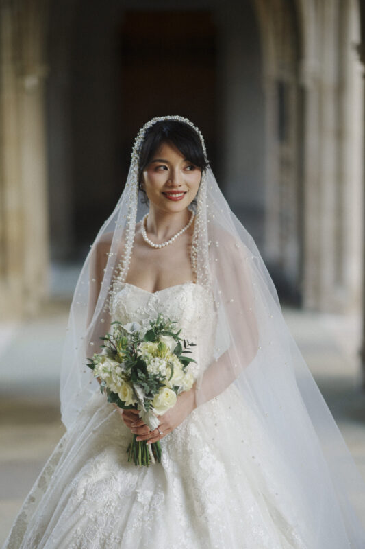 A bride stands in a corridor wearing a white wedding dress with a pearl-encrusted veil. She holds a bouquet of white and green flowers, smiling softly. The background features tall stone arches.