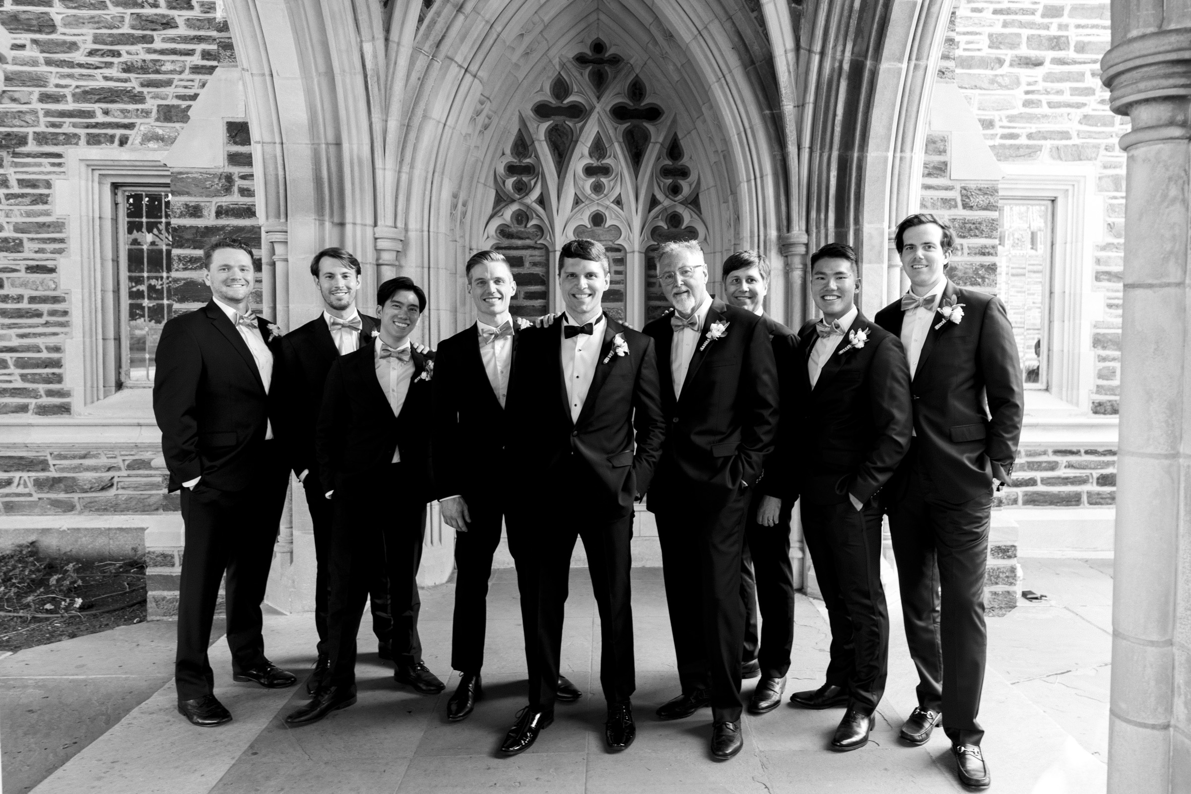 A group of nine men in suits and bow ties stand together, smiling in front of an ornate stone archway and wall. They appear to be part of a wedding party, with some wearing boutonnieres. The setting is elegant and formal.