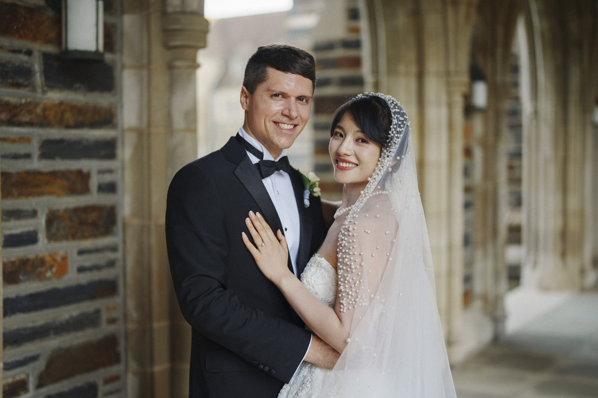 A bride and groom pose together in elegant wedding attire. The bride wears a white dress with a beaded veil, and the groom is in a black tuxedo. They stand in an arched corridor with stone walls, smiling at the camera.