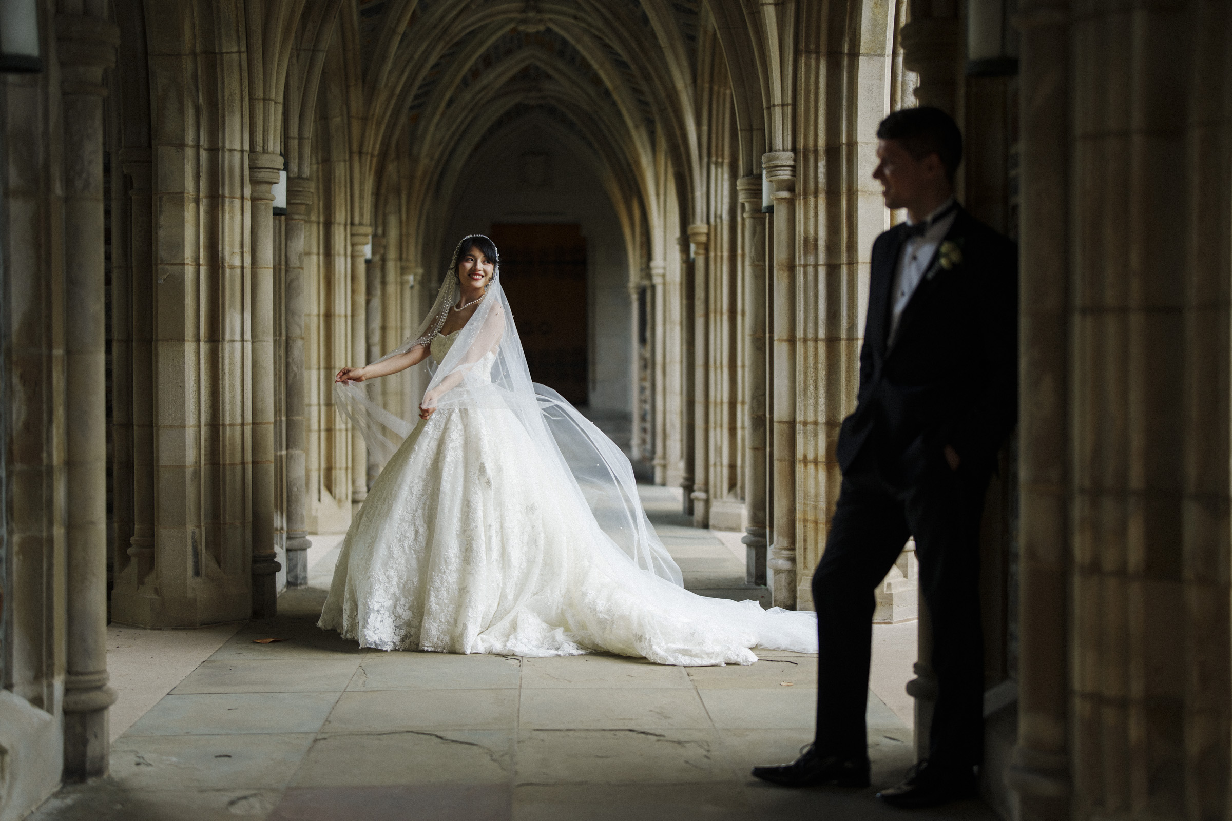 A bride in an elegant white gown and veil stands in a stone-arched corridor, smiling towards a groom in a black tuxedo who leans against a column. The setting has an old-world, romantic atmosphere.