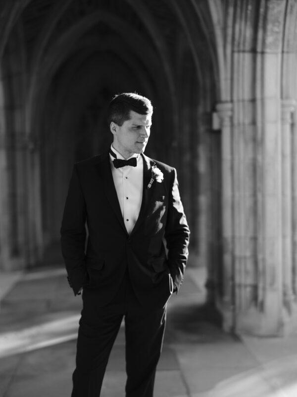 A man in a tuxedo stands in an arched, stone corridor, looking to the side. The setting is illuminated by soft, natural light highlighting the details of the stonework and his formal attire.