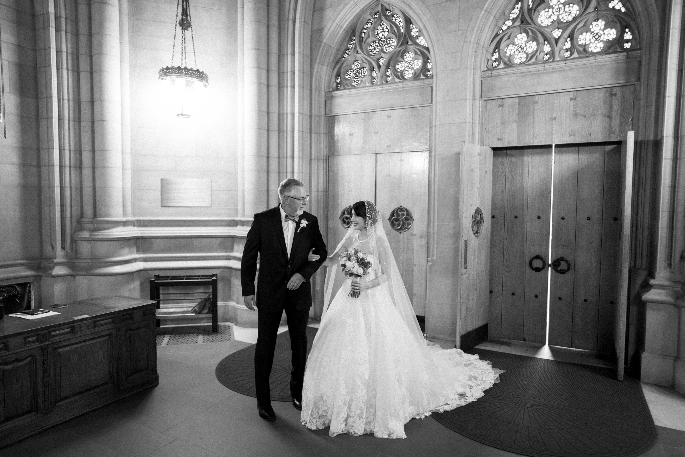 A bride in a white gown and veil holds a bouquet and walks arm-in-arm with an older man in a suit, likely her father, in a grand, ornate building with arched doorways and intricate stonework.