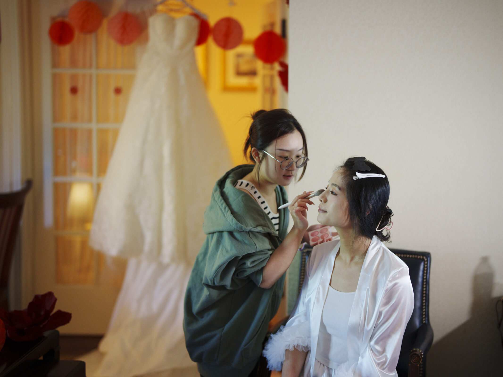 A makeup artist applies makeup to a woman wearing a white robe. In the background, a white wedding dress hangs on a door, and orange paper lanterns are visible. The setting appears to be a room with soft lighting.