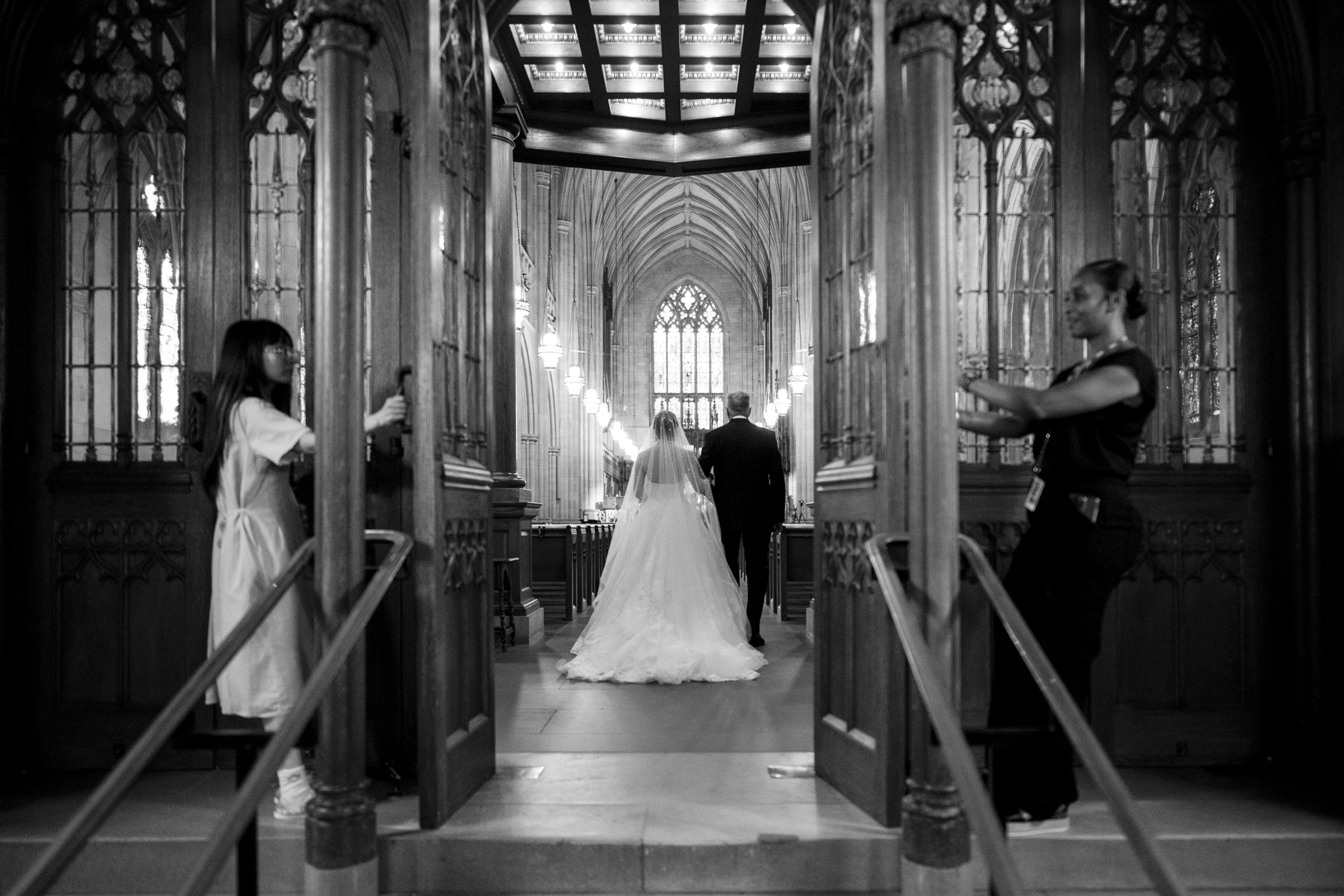 A bride and groom walk down the aisle of a grand church with high vaulted ceilings, as two attendants open large wooden doors behind them. The setting is elegant and illuminated with soft lighting.