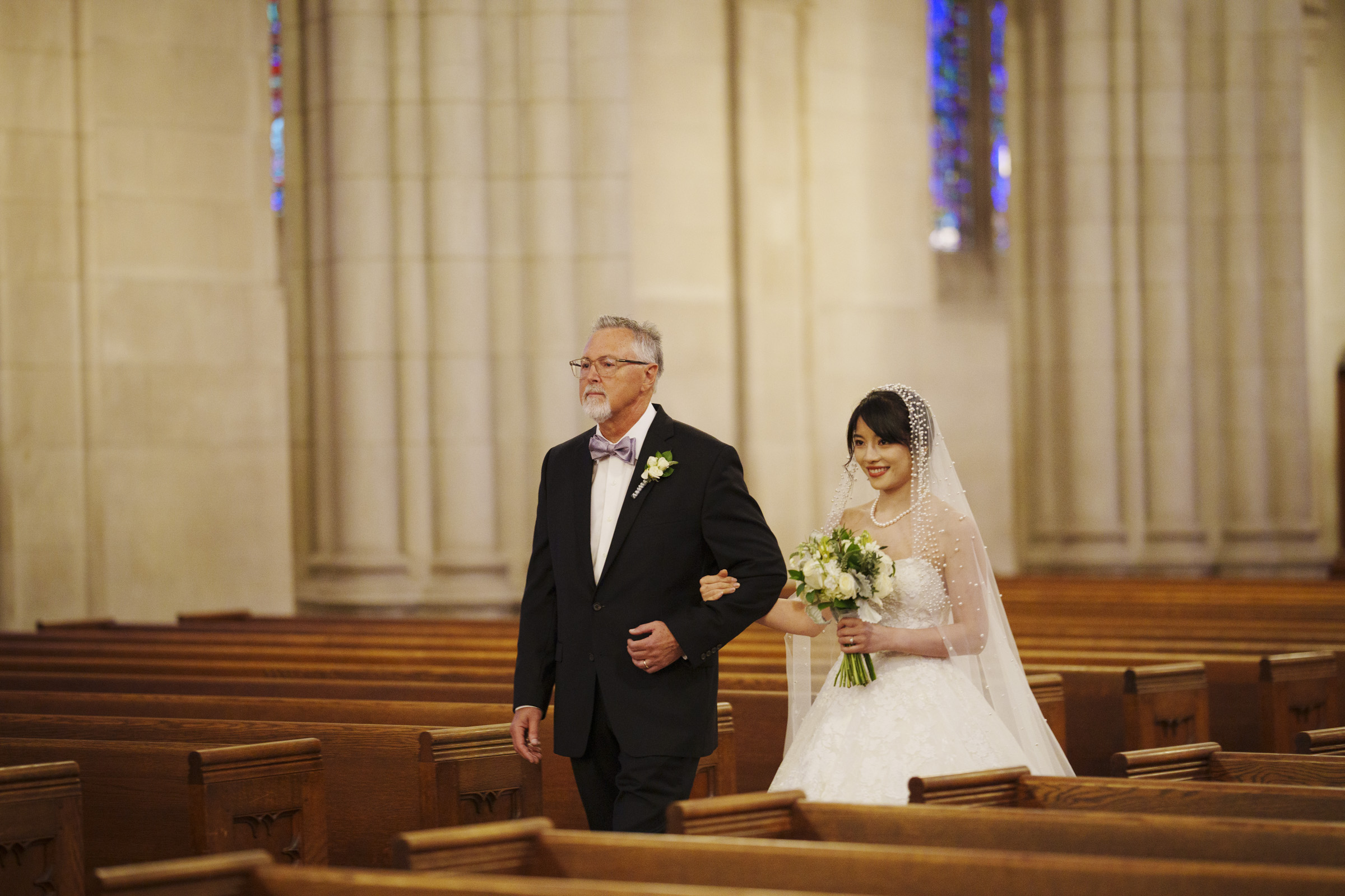 A bride in a white wedding dress and veil holds a bouquet while walking down the aisle with an older man in a suit and bow tie, inside a church with wooden pews and stained glass windows.