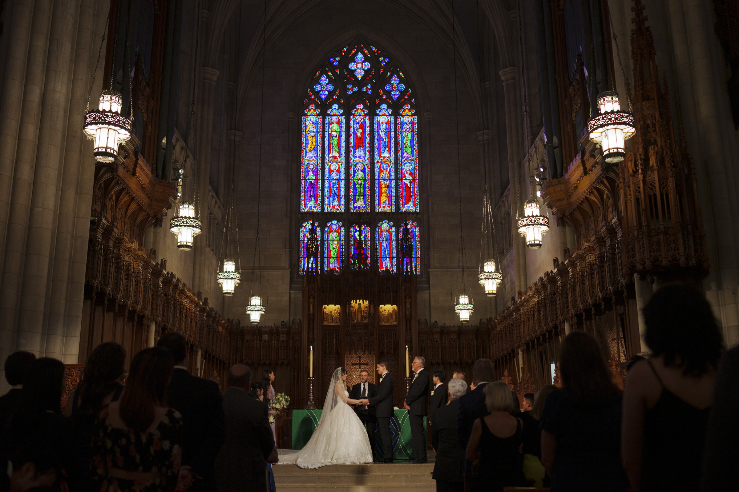 A wedding ceremony taking place inside a grand cathedral with high vaulted ceilings and ornate stained glass windows. The couple stands at the altar, surrounded by guests seated in pews. The setting is dimly lit with decorative hanging lamps.