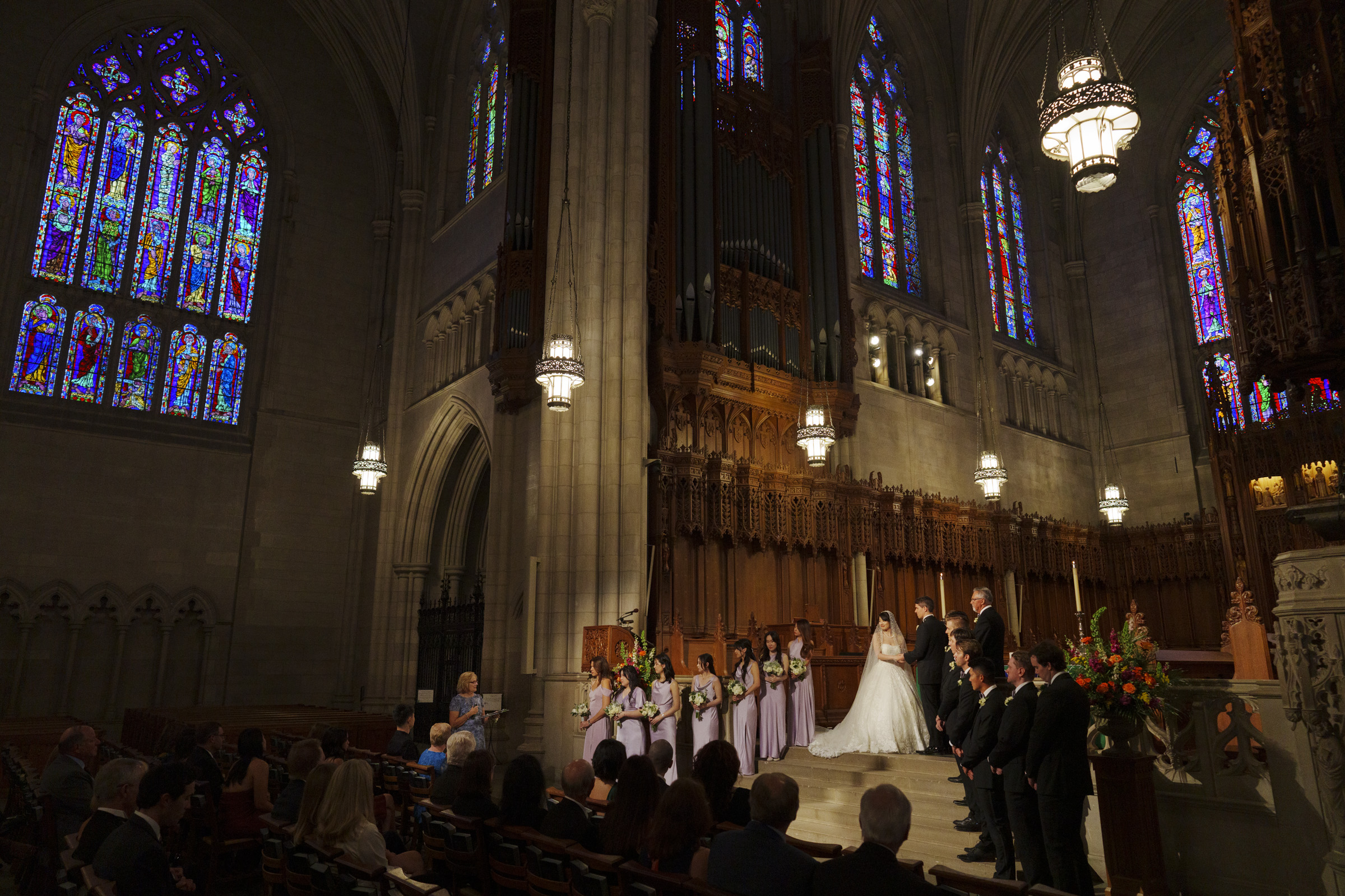 A wedding ceremony inside a grand church with stained glass windows. The bride and groom stand at the altar, surrounded by a bridal party in lavender dresses and formal suits. The audience is seated in pews, watching the ceremony.