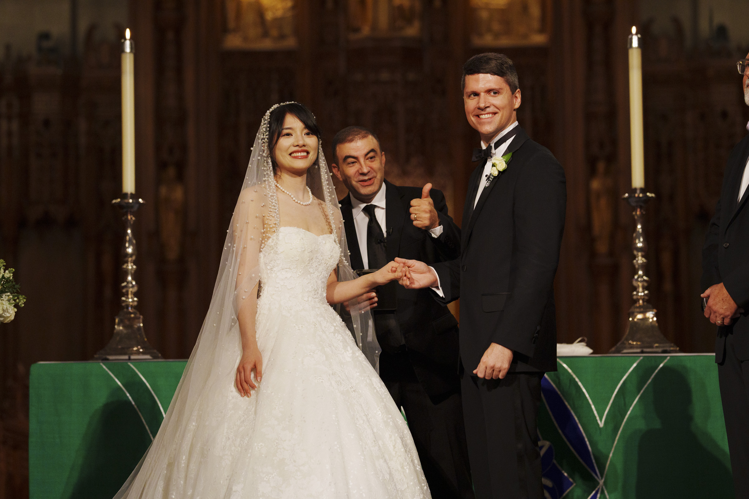 A bride and groom stand smiling, holding hands. The bride wears a white gown and veil. The groom is in a black tuxedo. A man beside them, also in a suit, gives a thumbs-up. Theyre standing in front of an altar with two tall candles.