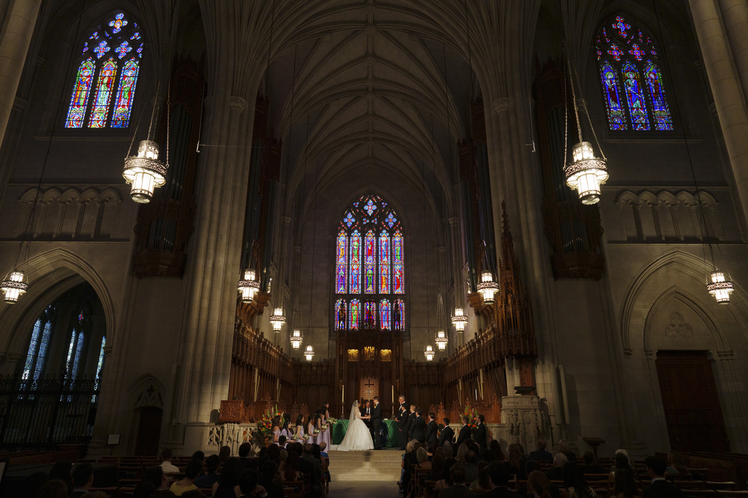 A grand cathedral interior with high vaulted ceilings and colorful stained glass windows. A wedding ceremony is taking place, with the couple standing before an altar and guests seated in pews. The space is dimly lit with hanging chandeliers.