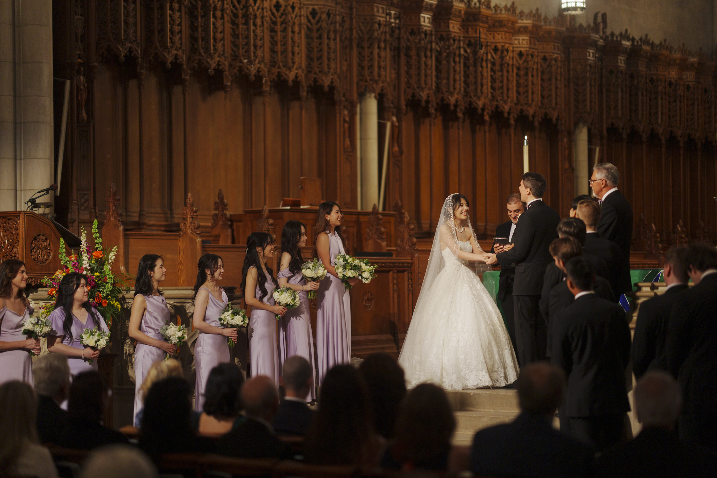 A bride and groom stand at an ornate altar during a wedding ceremony. The bride is in a white gown, and the groom is in a suit. Several bridesmaids in lavender dresses and groomsmen are present. An audience is seated in the foreground.