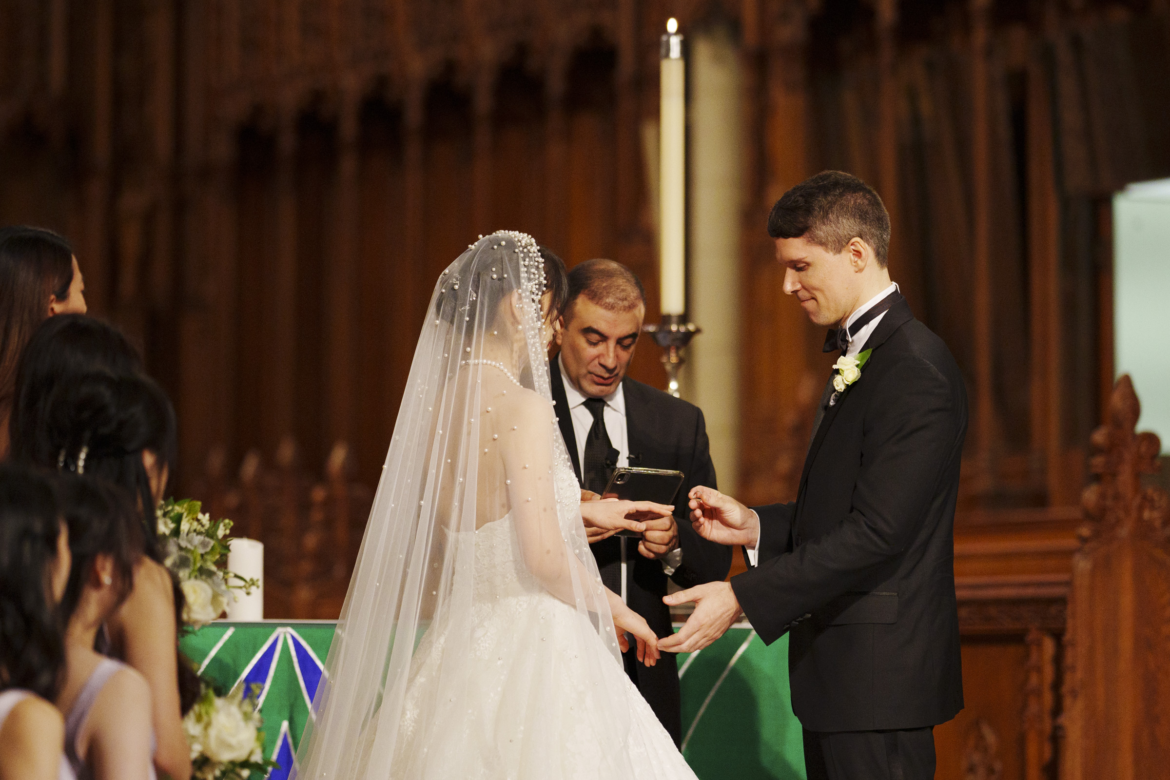 A couple stands at an altar during a wedding ceremony. The bride is wearing a white dress with a veil, and the groom is in a black suit. A man in a black robe is officiating the ceremony, holding a book. A large candle is visible behind them.