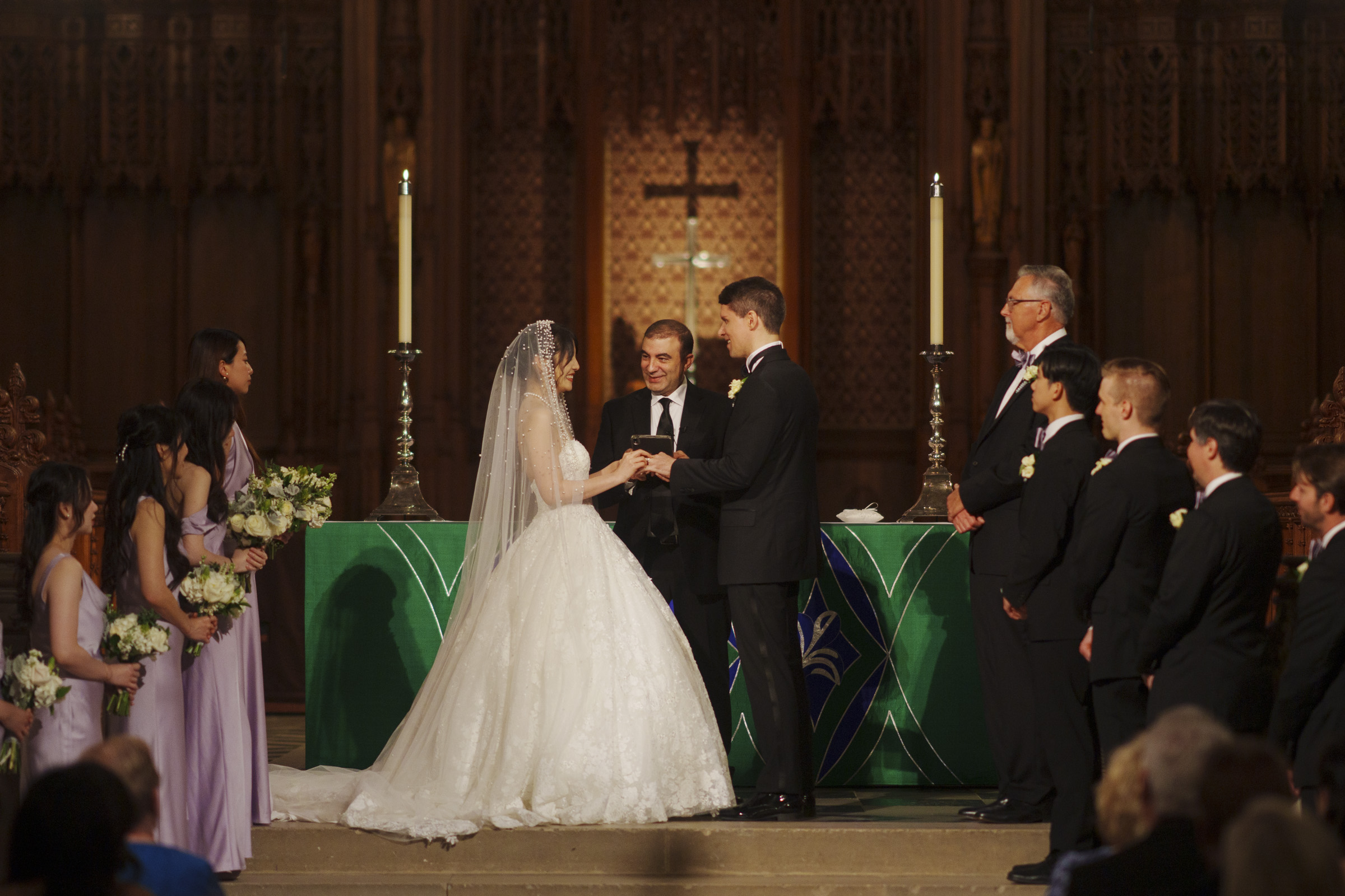 A bride and groom stand at the altar during a wedding ceremony in a church. The bride wears a white gown and veil, and the groom is in a black suit. Bridesmaids in lavender dresses and groomsmen in black suits are positioned on either side.