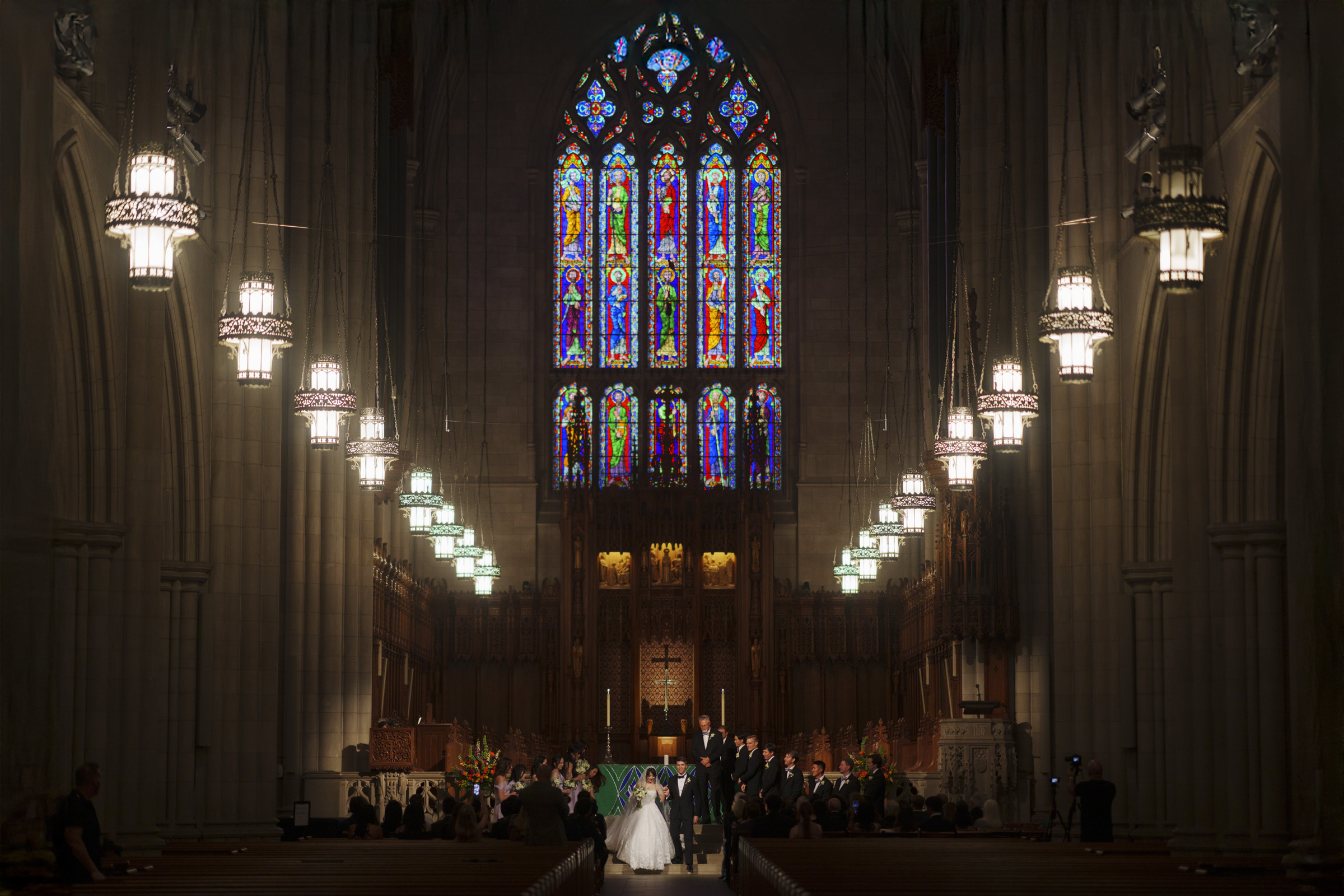 A couple stands at the altar in a grand cathedral with high ceilings. Stained glass windows above cast colorful light into the dimly lit space. Large chandeliers and wooden pews line the aisle, creating an elegant atmosphere for the wedding ceremony.