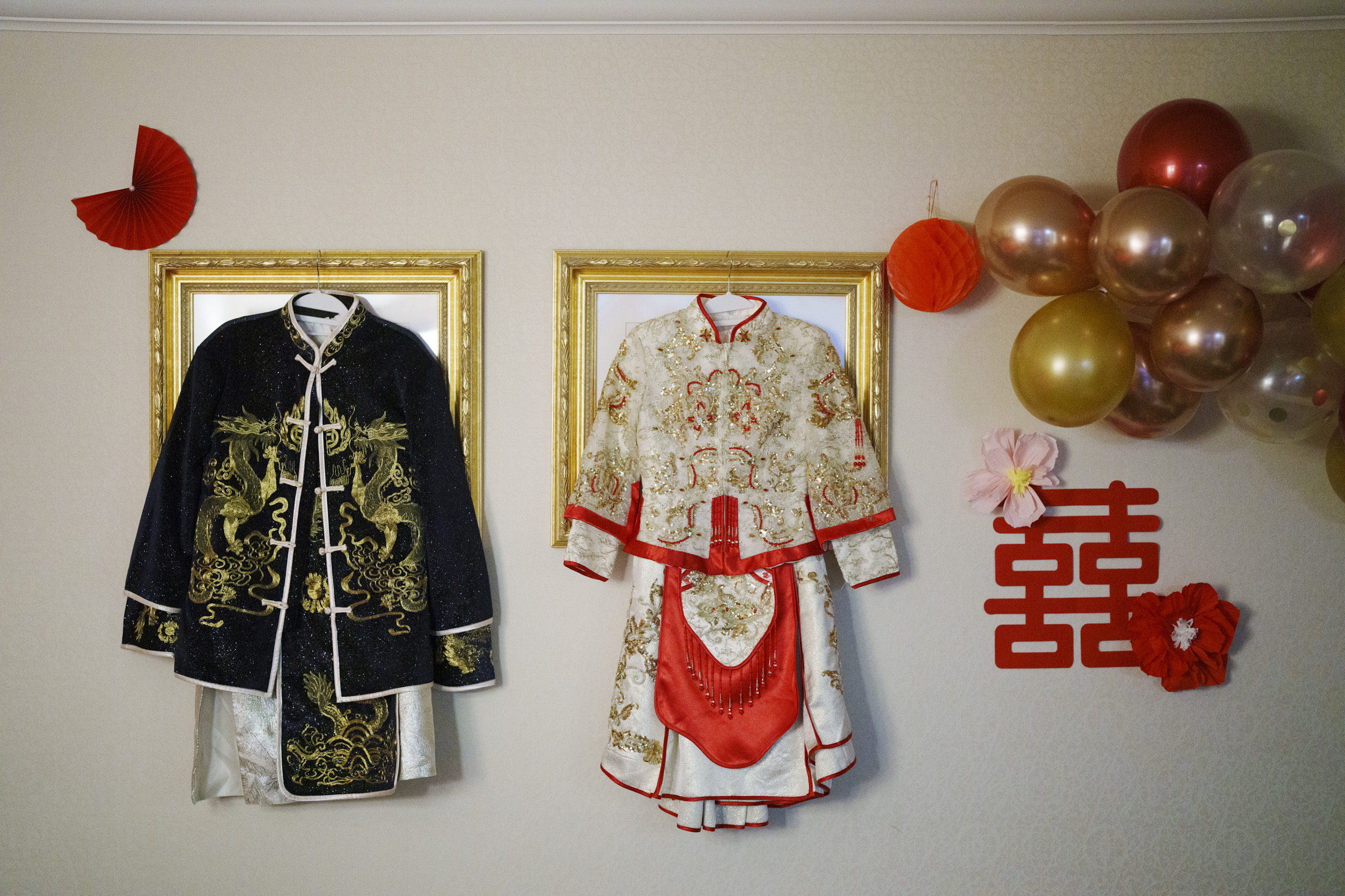 Traditional embroidered wedding attire displayed on a wall with gold frames. The clothing includes a dark jacket with gold dragons and a white dress with red accents. Decorated with red and gold balloons, red fans, and a double happiness symbol.