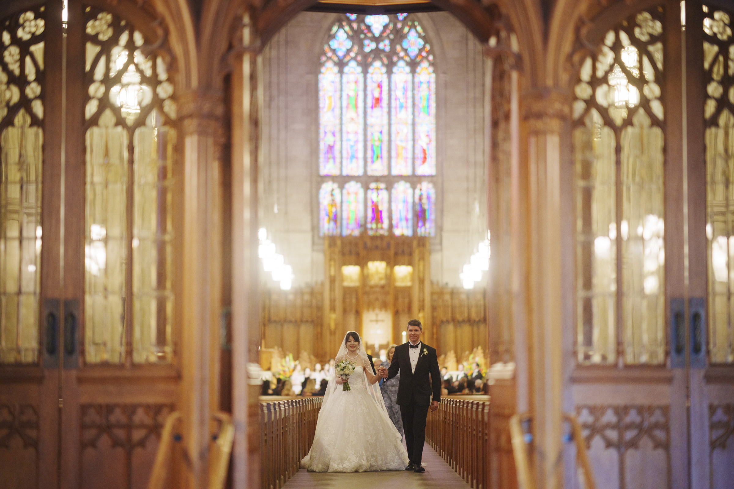 A bride and groom walk down the aisle in a grand cathedral. The interior features ornate stained glass windows and wooden arches, creating a majestic atmosphere. The couple is dressed formally, with the bride in a white gown and the groom in a dark suit.